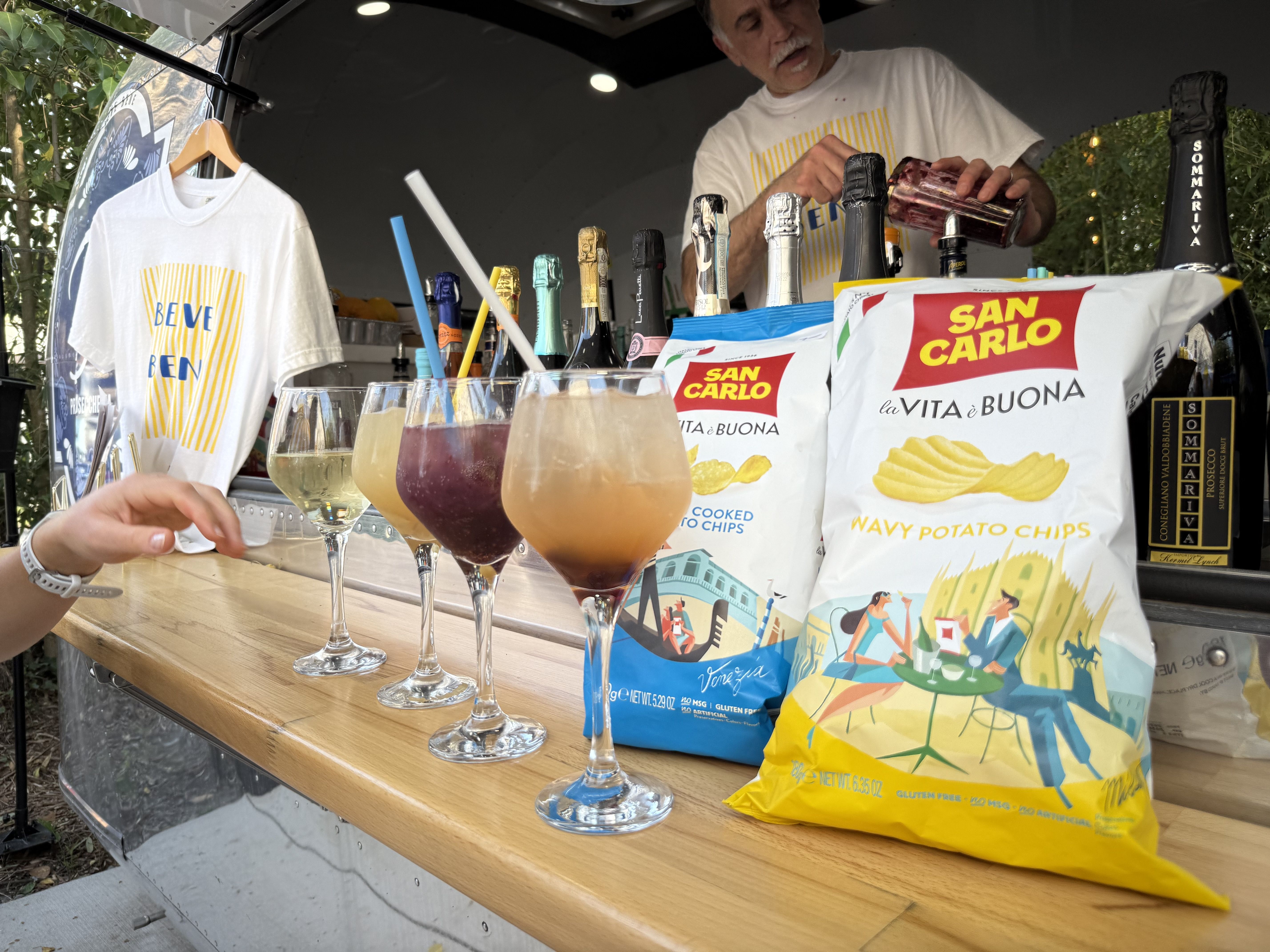 Mobile bar on a truck with assorted cocktails in stem glasses, a bartender in a white shirt behind the counter, and large San Carlo potato chip bags on a wooden foreground counter.