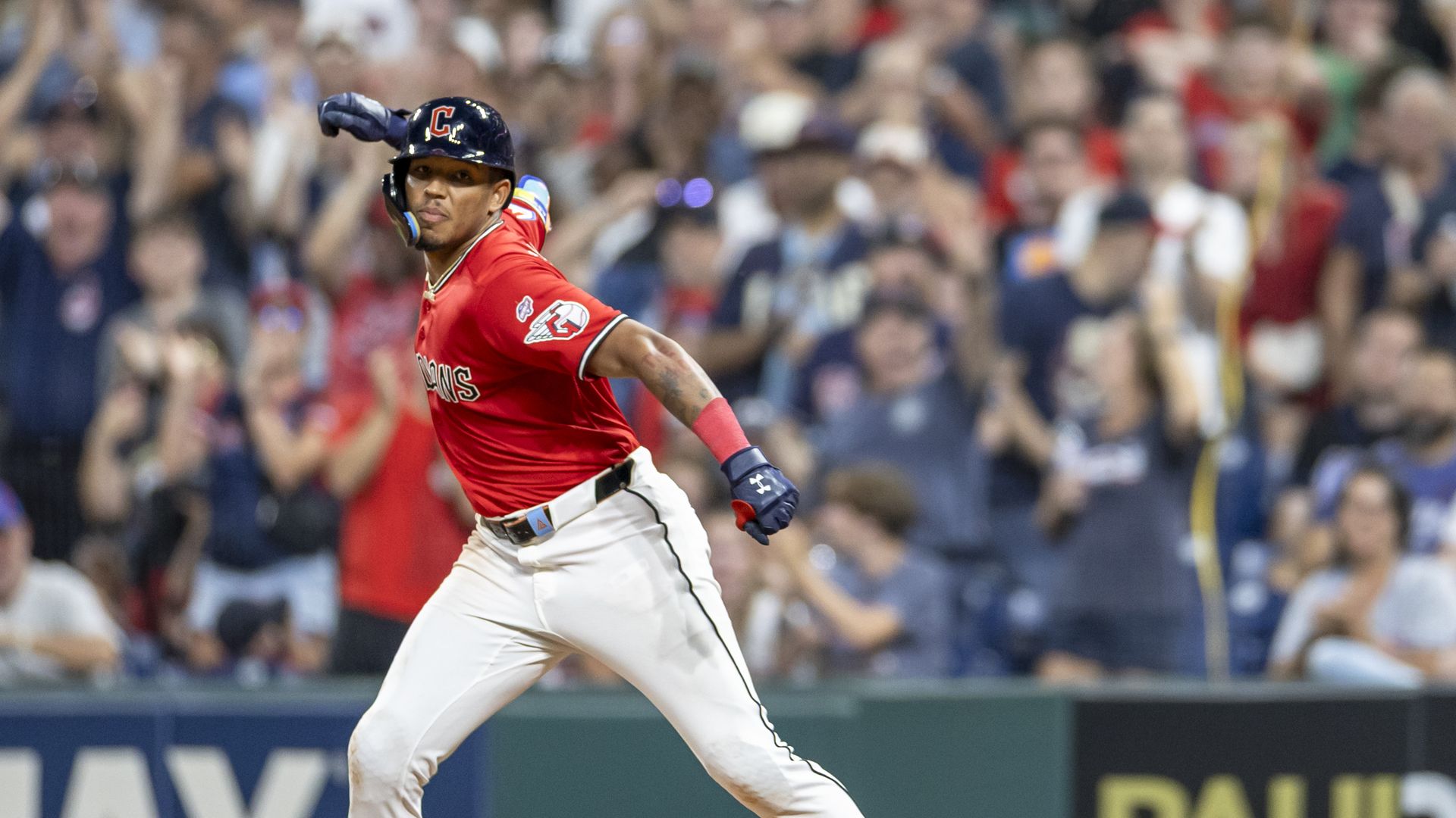 Baseball player in a red jersey and white pants runs on the field, wearing a black helmet with a red C and blue gloves, with a cheering crowd blurred in the background.