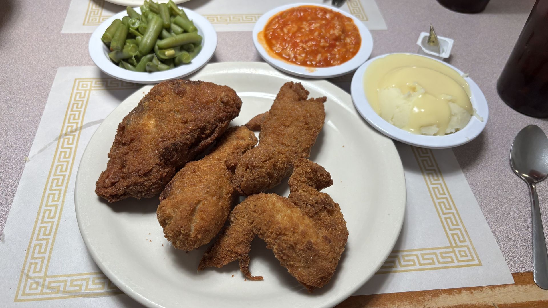 Plate of crispy fried chicken pieces with sides of green beans, tomato-based sauce, and mashed potatoes with gravy, set on a beige patterned placemat at a dining table.