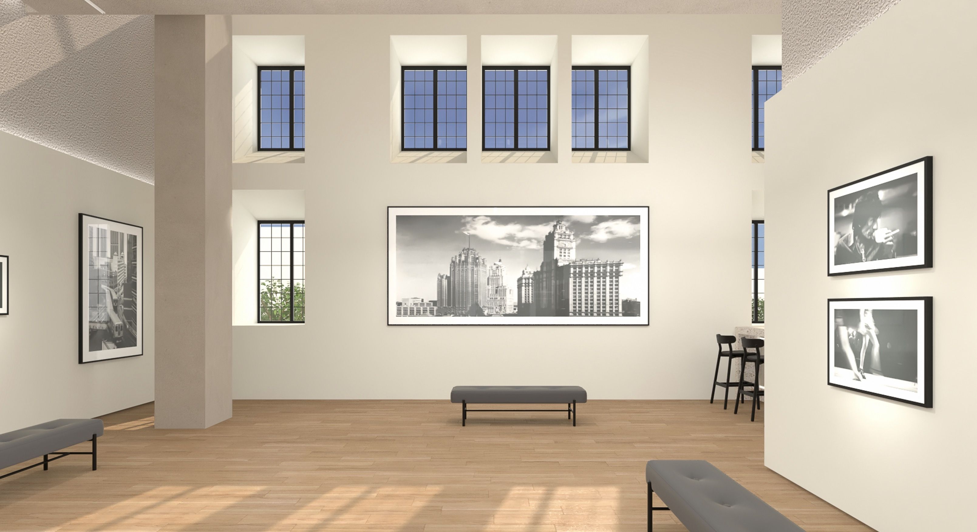 Bright modern art gallery with beige walls and light wood floors. A large black-and-white cityscape photo hangs center; benches line the room. Framed photos adorn side walls near tall grid windows.