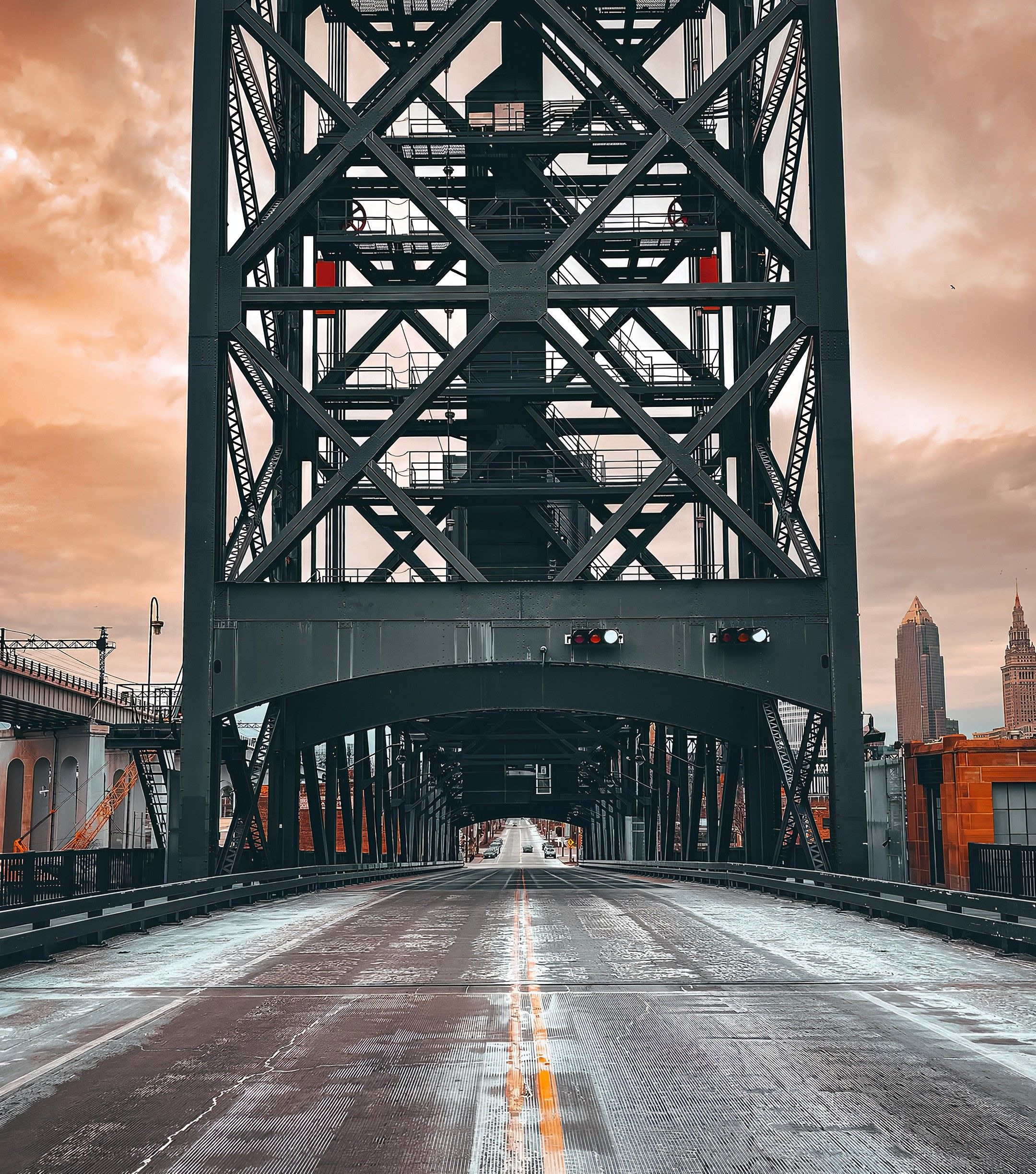 Empty gray metal truss bridge with a road centerline, under a cloudy orange sky, cityscape with tall buildings visible in the background.