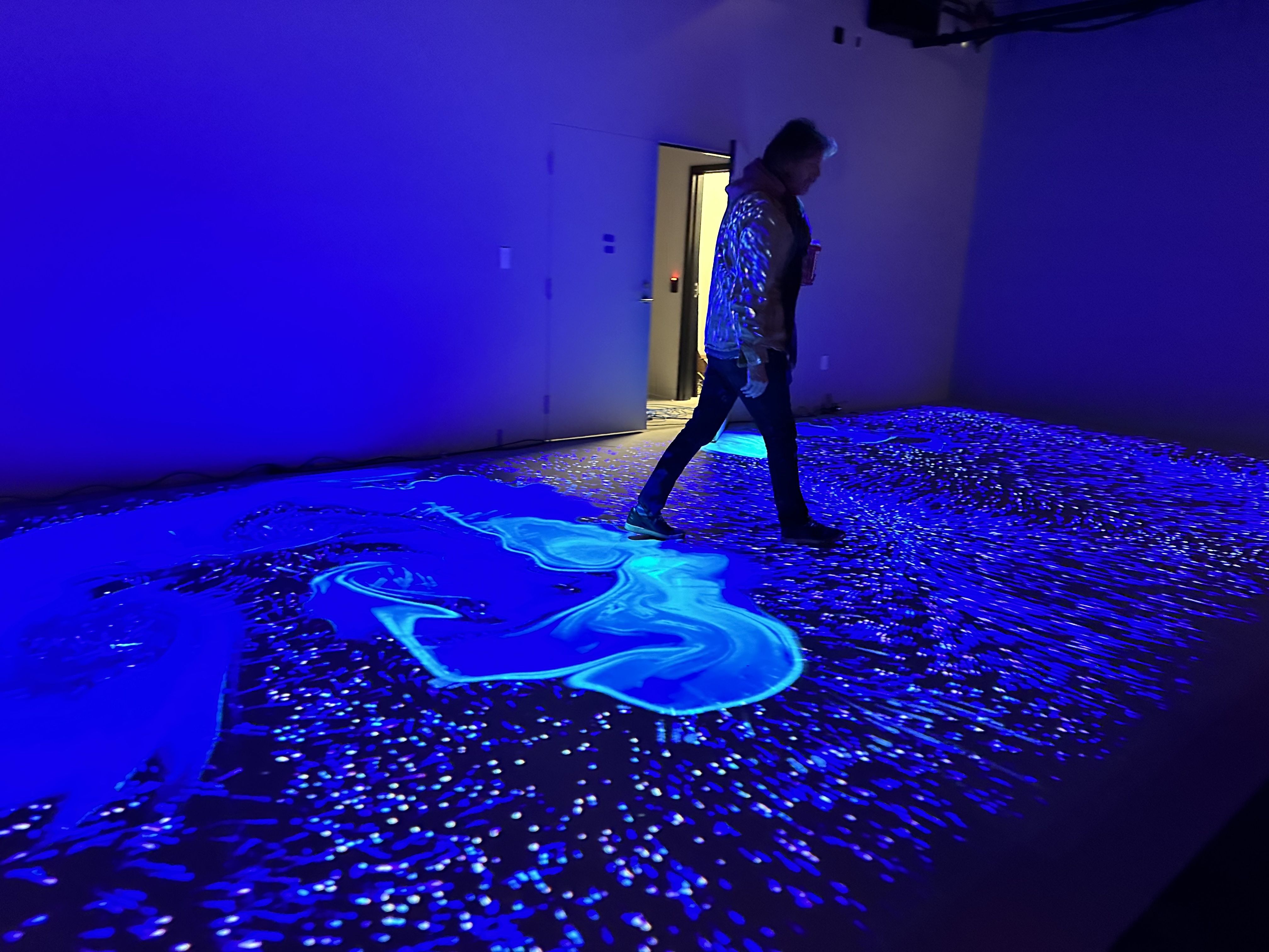 A man walks through a dark room with blue lights projected onto the floor that move whenever someone steps on them/the floor at WNDR Museum.