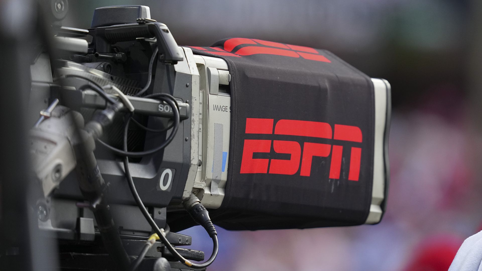 Close-up of a professional video camera with a black cover displaying the red ESPN logo, set against a blurred background.