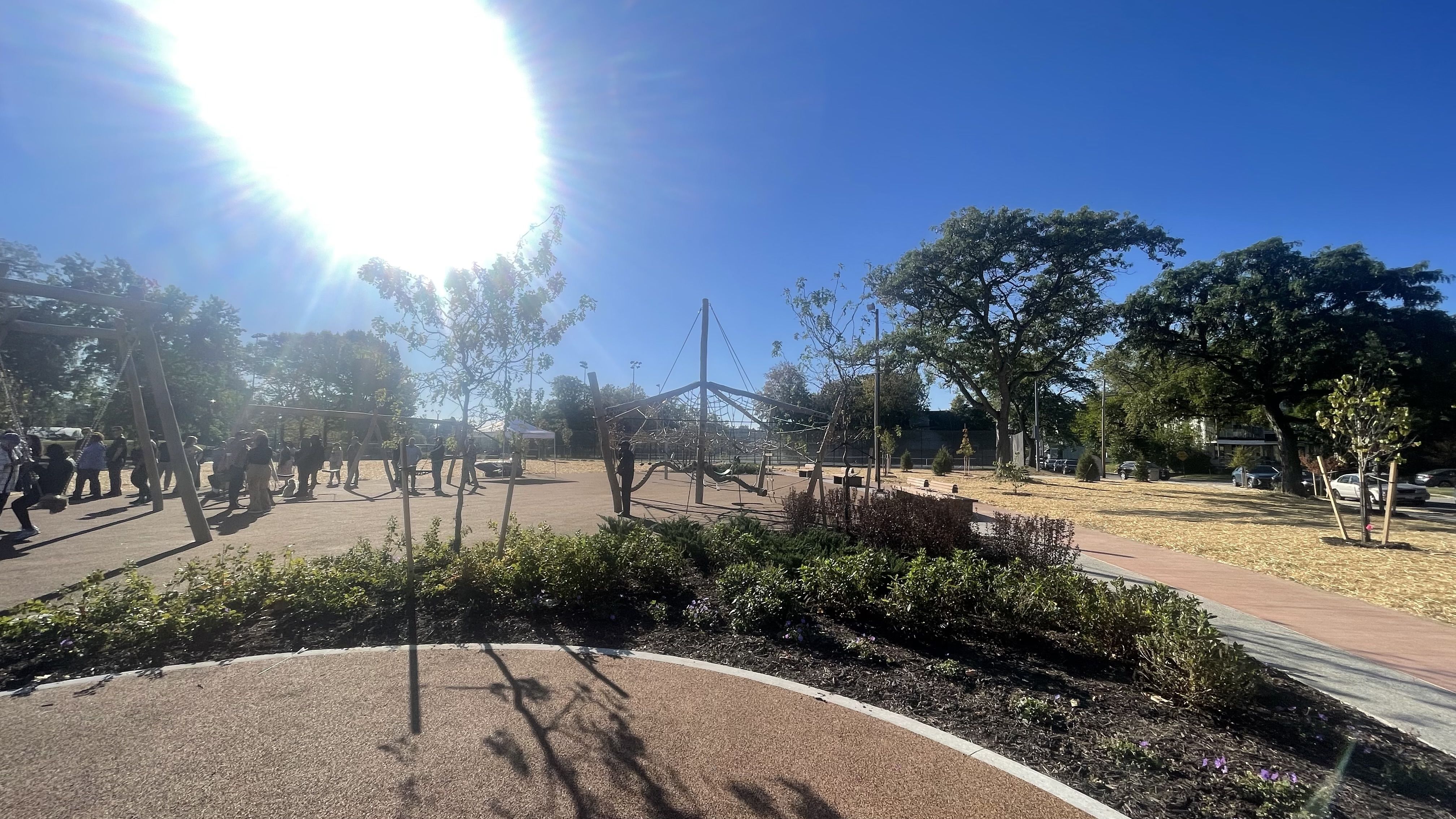 Bright sunny day at a playground with swings, climbing equipment, people gathered, green bushes, trees, clear blue sky, and a sun glare on the left side.