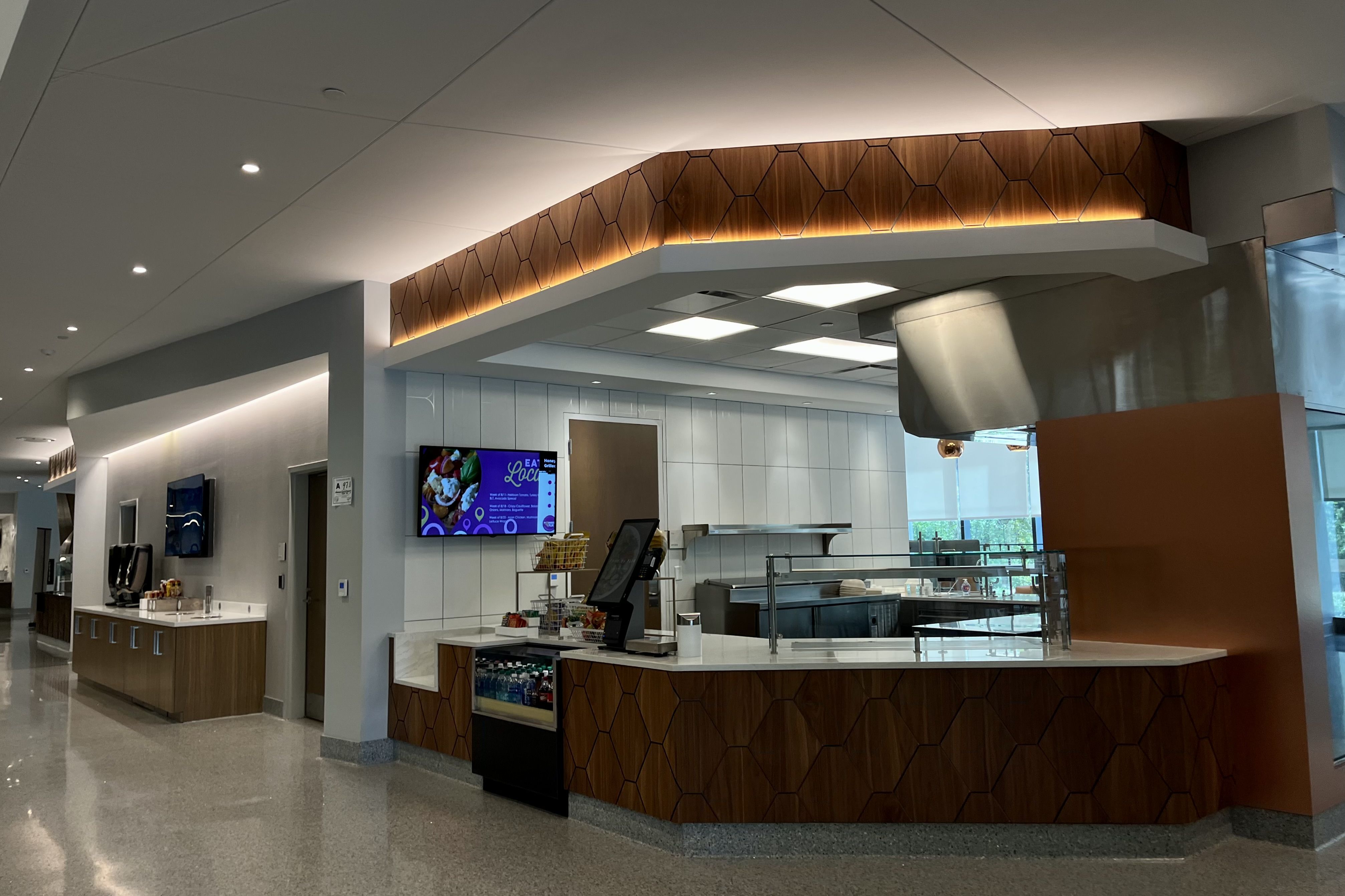 Modern indoor cafeteria with wooden hexagonal paneling, white countertops, a soda machine, digital menu screen, and clean tile floors under soft ceiling lights.