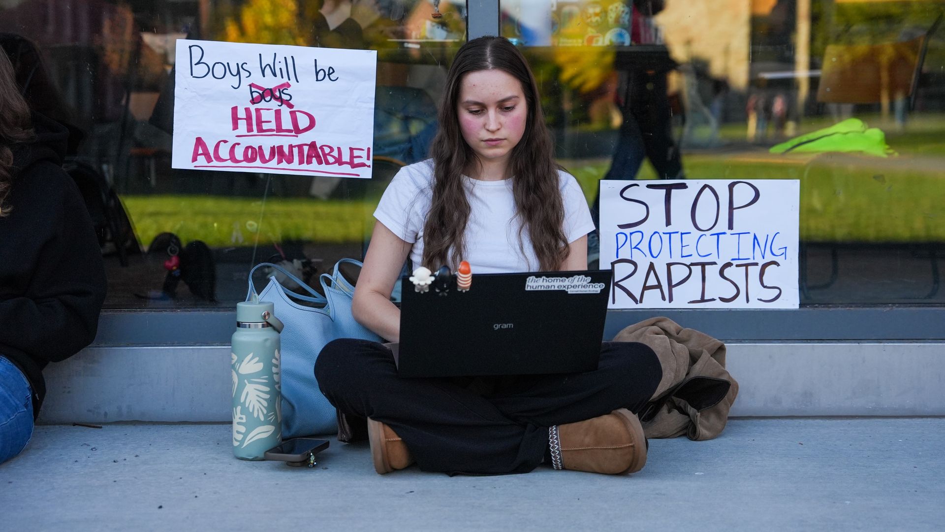 Young woman sits cross-legged by a window with a laptop. Left protest sign reads Boys Will Be Boys, crossed out, with HELD ACCOUNTABLE in pink; right sign says STOP PROTECTING RAPISTS.