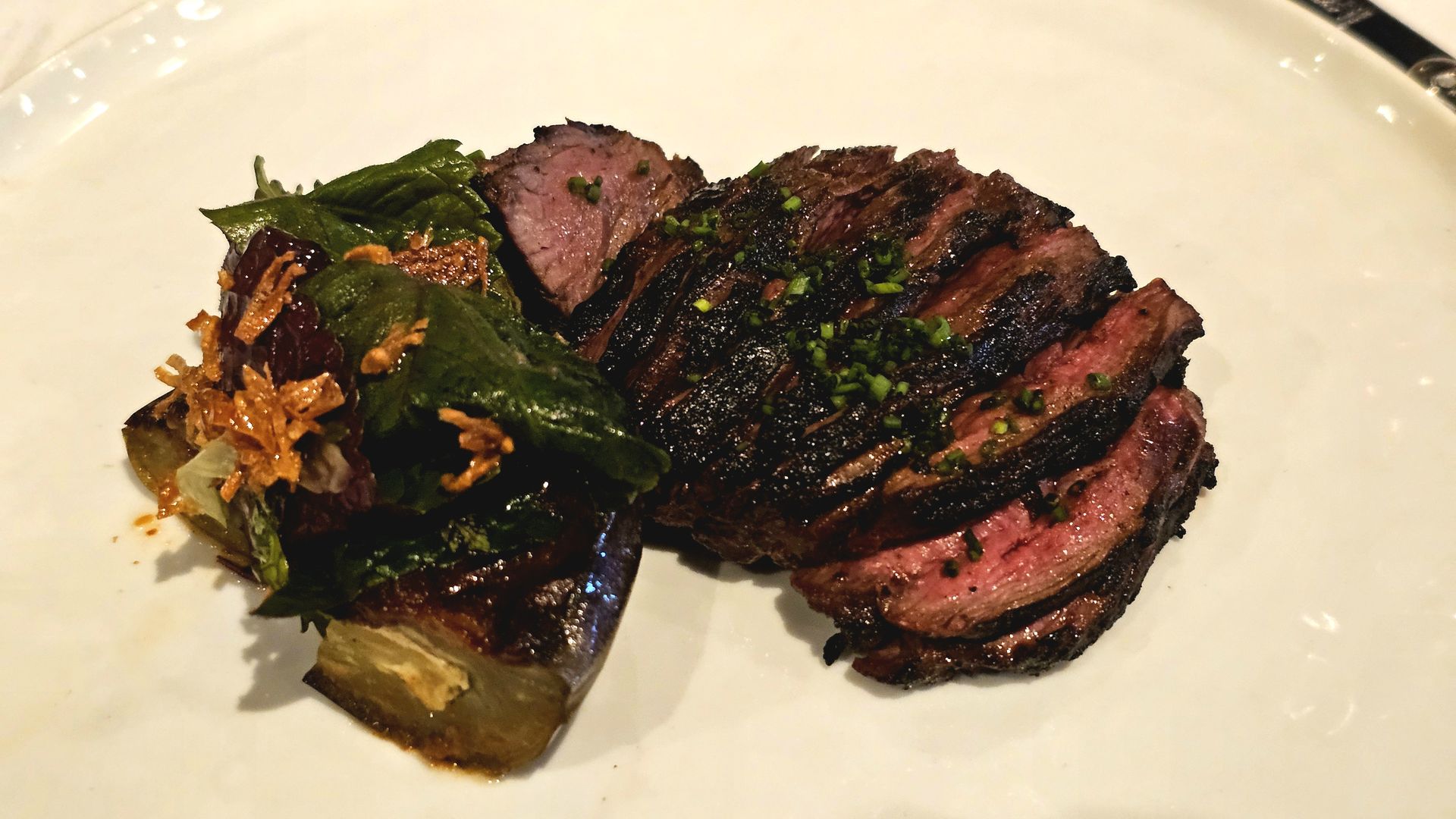 Plate of sliced, medium-rare steak with a blackened crust and pink center, garnished with chopped green herbs, on a white plate with greens and crispy fried onions on the side.