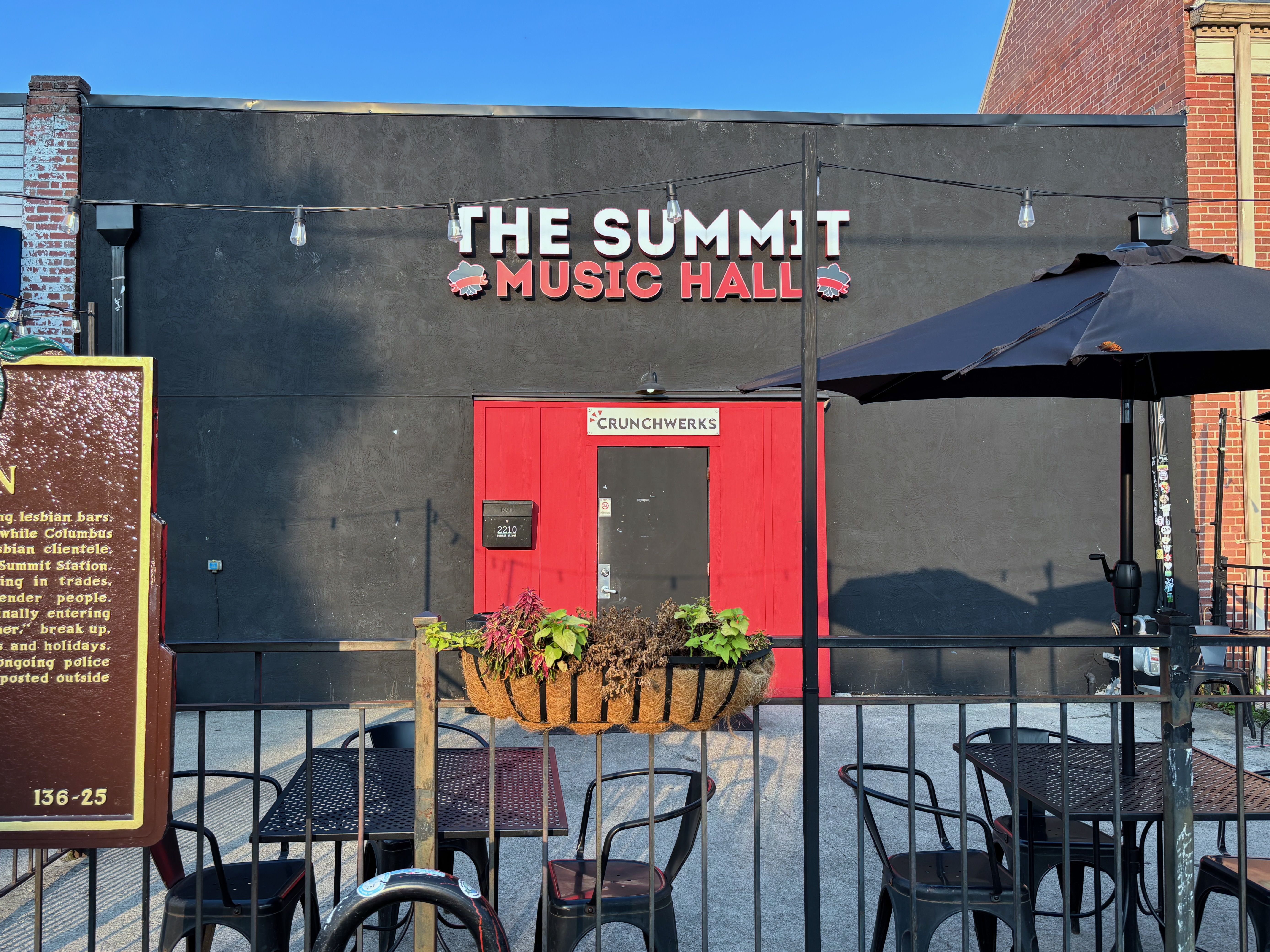 The exterior of the Summit Music Hall, a black building with a red door that says "Crunchwerks" above it, with patio tables and an umbrella in the foreground.