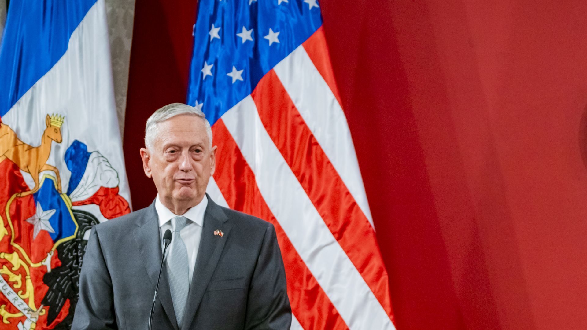 Jim Mattis purses his lips standing before a red background and two flags