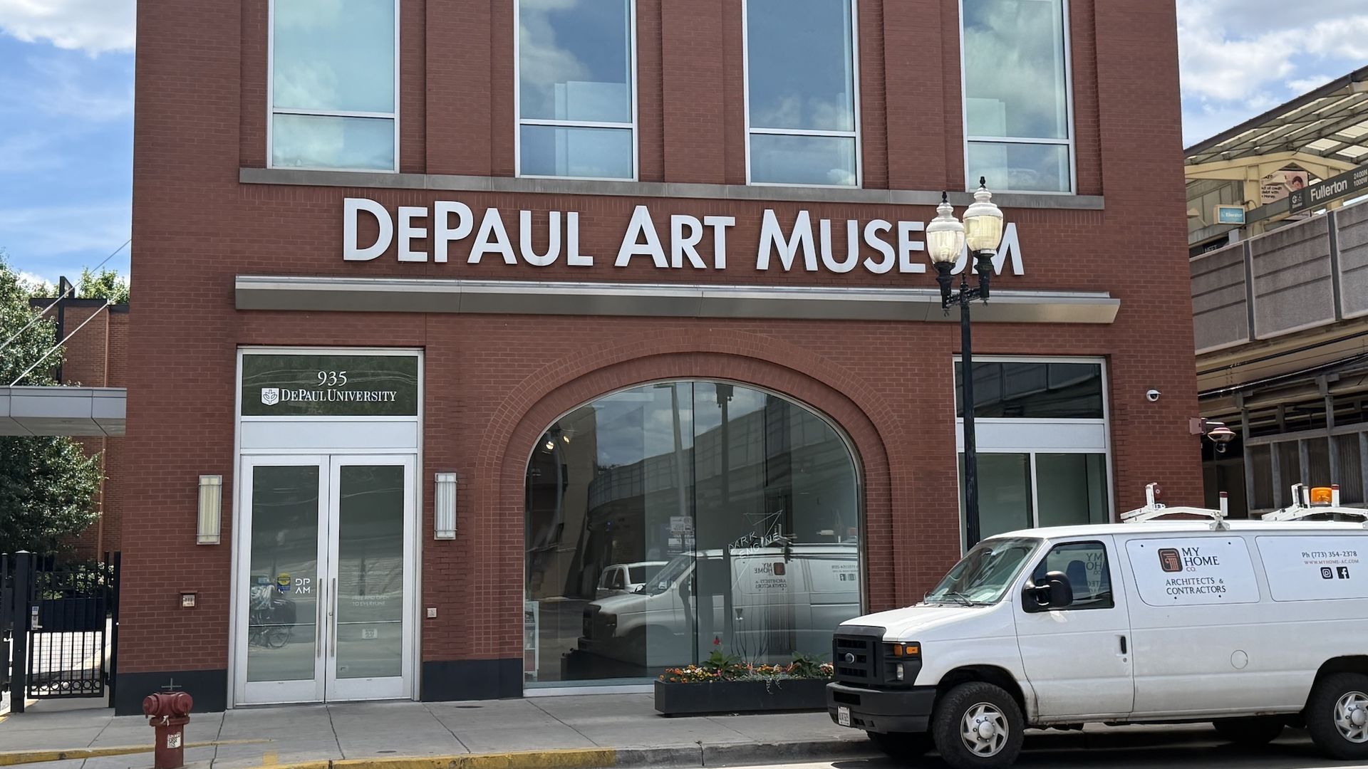 Brick DePaul Art Museum building with a large white sign reading "DEPAUL ART MUSEUM", tall windows, a vintage street lamp, and a white van parked along the sidewalk under a blue sky.