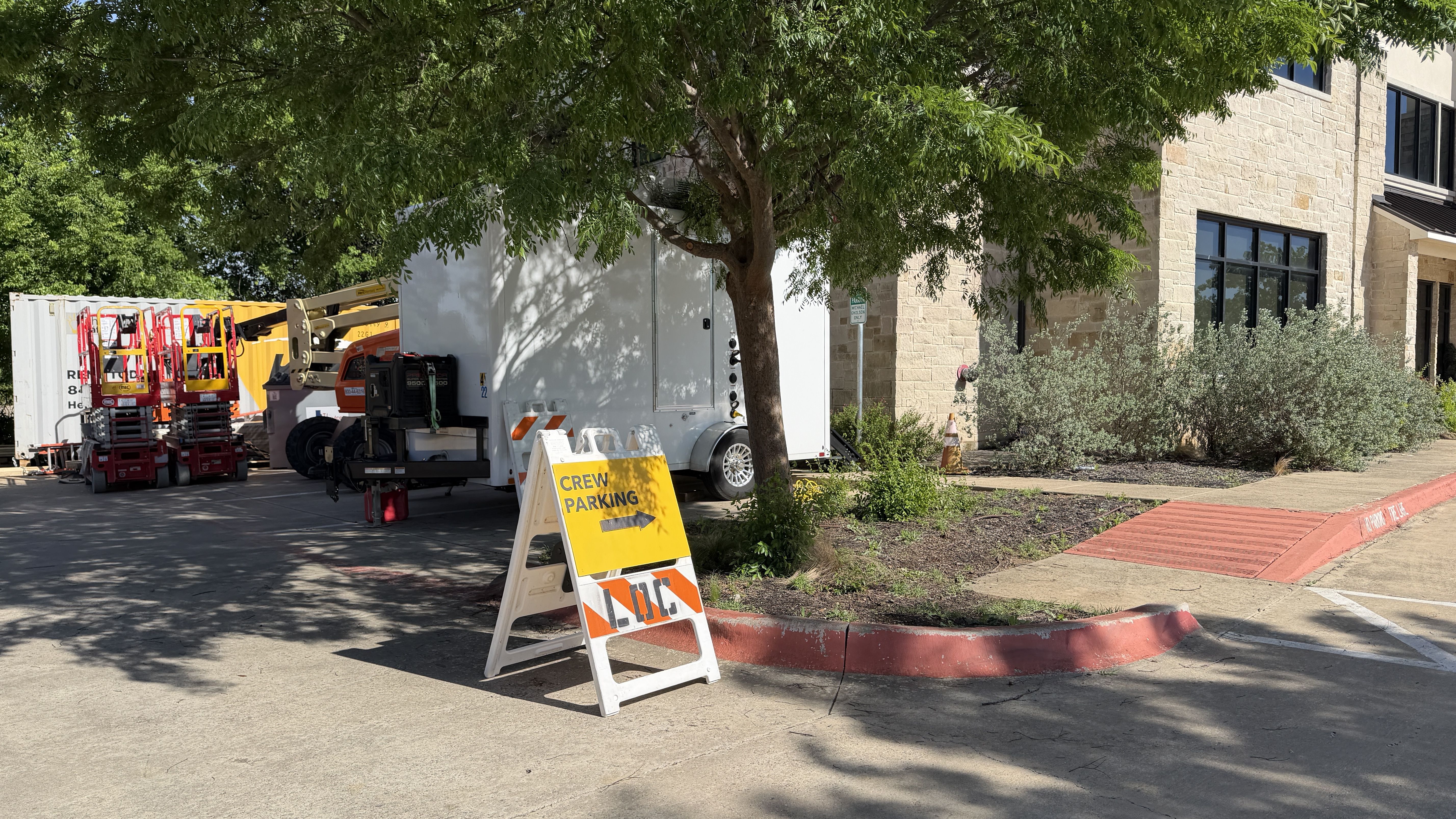 Construction area beside a beige stone building: a white trailer, red/orange aerial lifts, and a bright yellow "Crew Parking" sign under a leafy green tree.