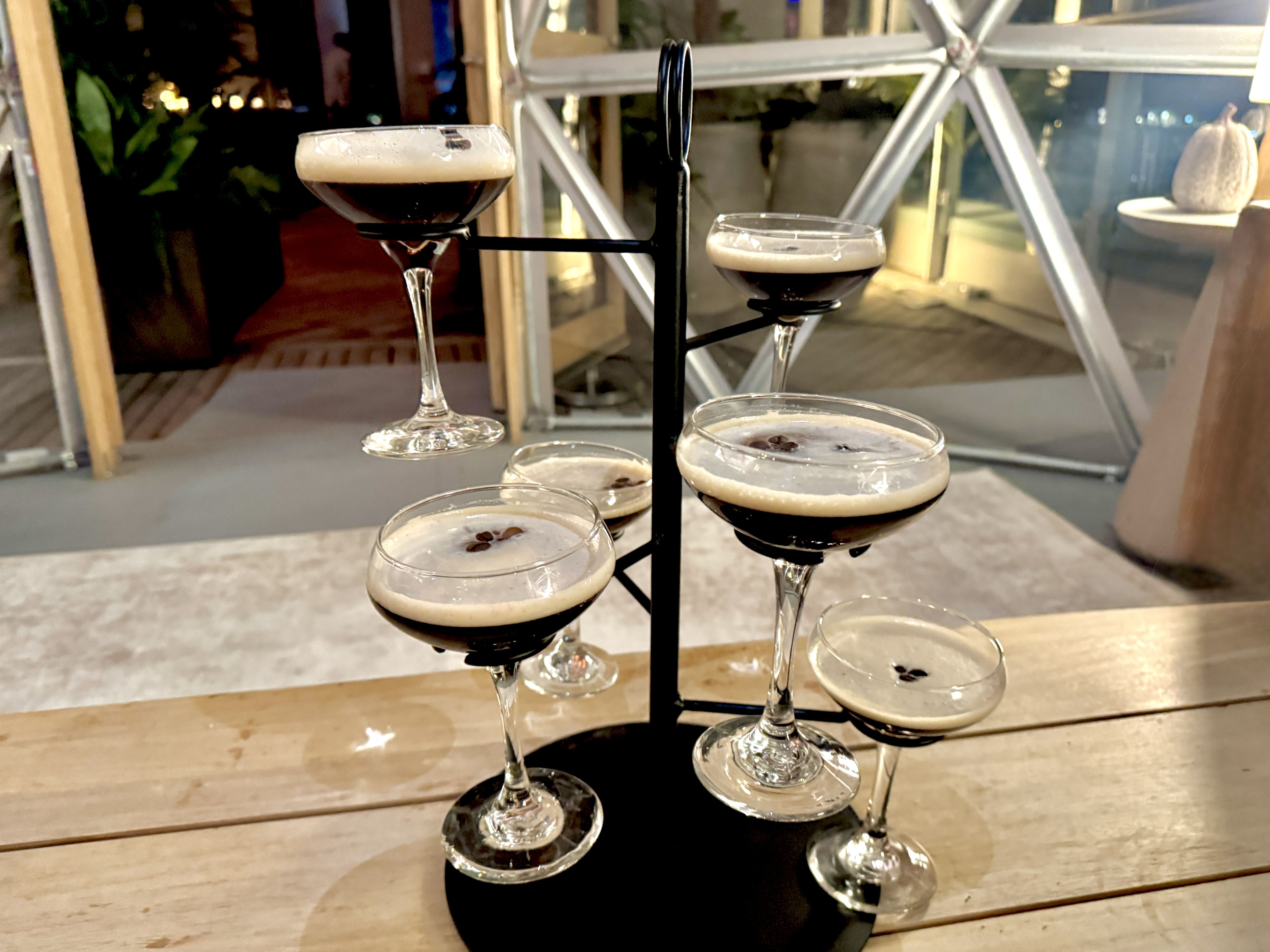 Six espresso martinis in coupe glasses arranged on a multi-level black metal stand on a wooden table inside a softly lit venue with glass walls.