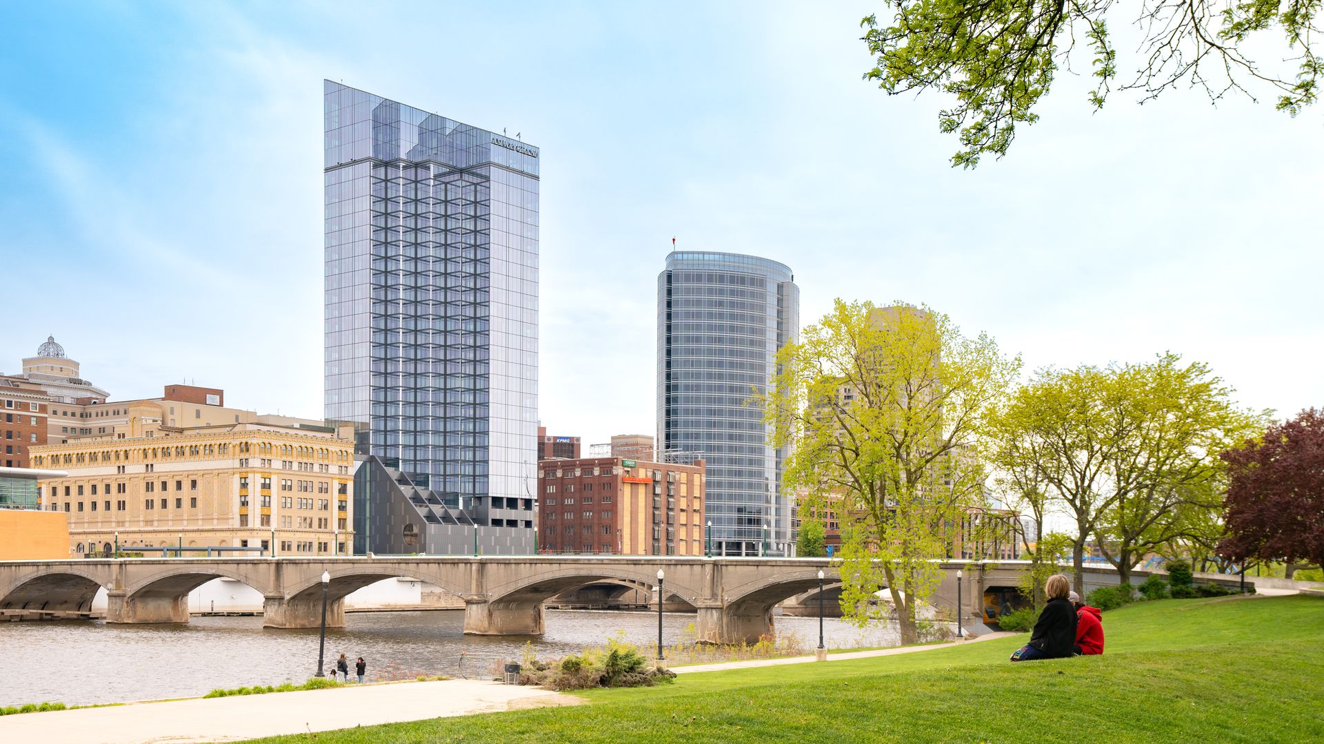 Buildings rising up behind a bridge over a river. A person sits watching the river on a grassy hill.