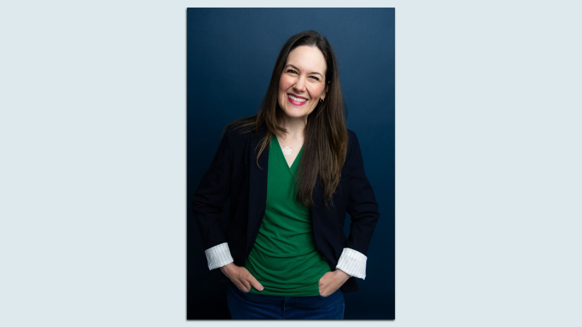 Smiling person with long brown hair, wearing a green shirt, dark blazer with rolled striped cuffs, and jeans, standing with hands in pockets against a dark blue background.