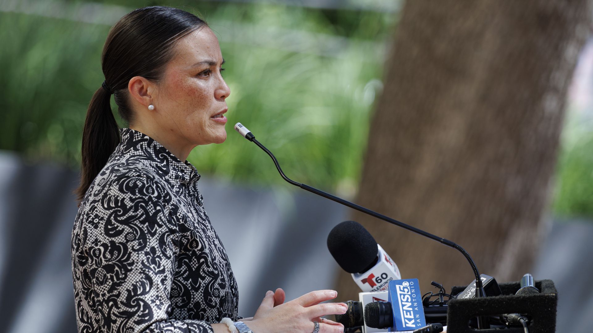 Mayor Gina Ortiz Jones is seen from the side at a press conference, standing at a podium with microphones that are labeled with local news station logos. A blurred background behind her shows a tree and grass.