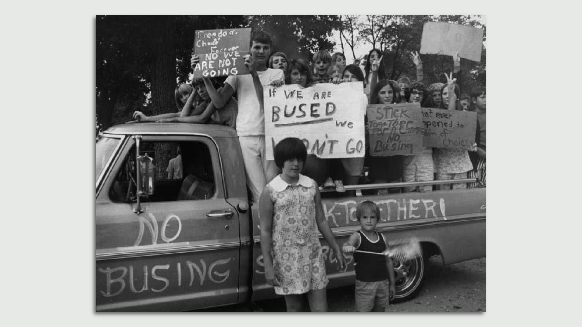 White children stand in the bed of a crappy pickup holding anti-school busing signs. 