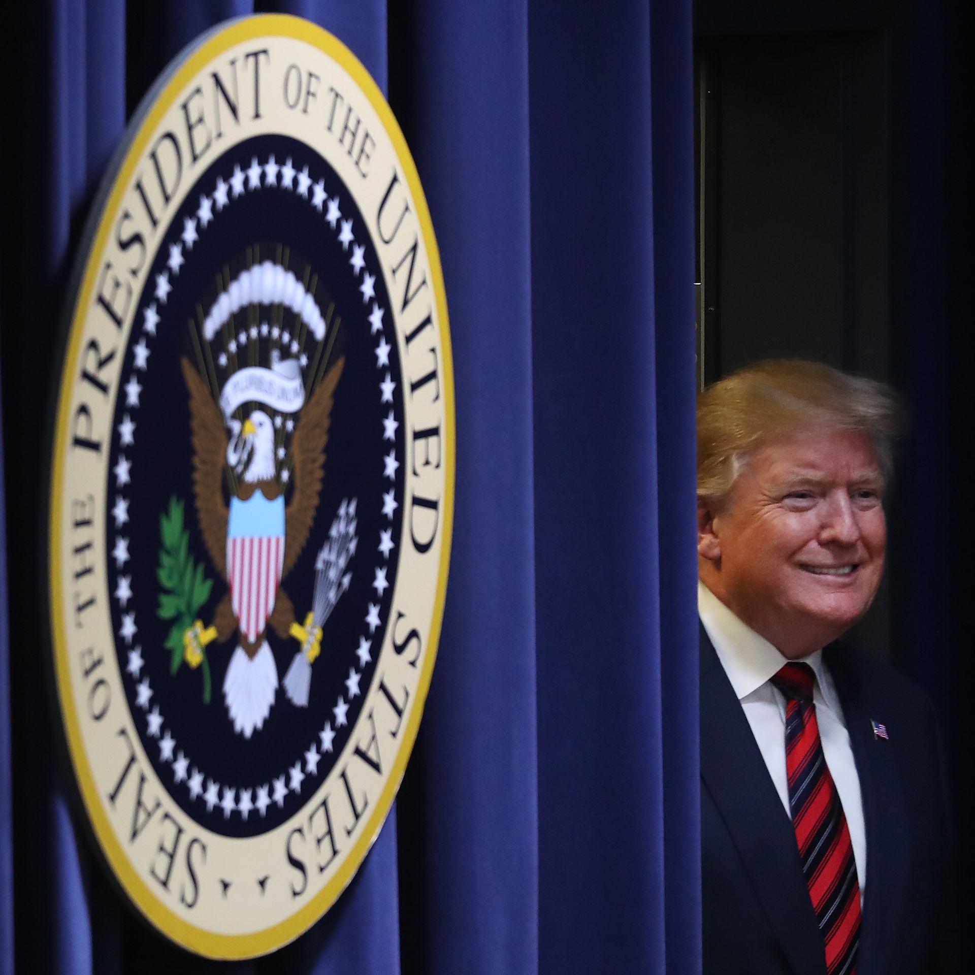 In this image, Trump smiles next to a heavy blue curtain next to the presidential seal. 