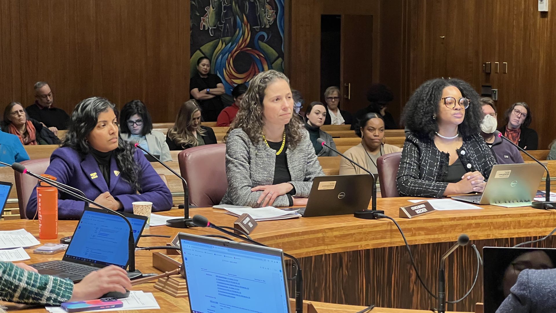 Three women in suits sit at a city council dais during a public meeting