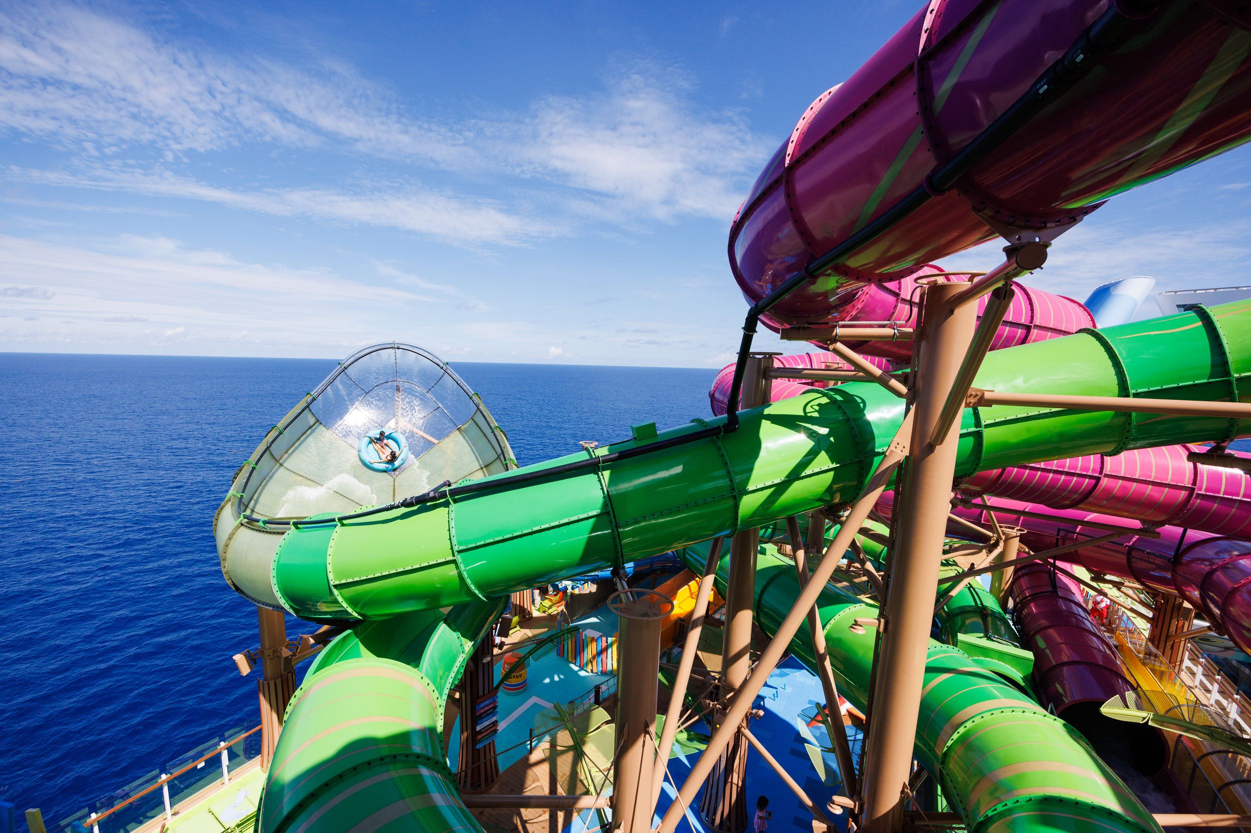 Colorful green and purple water slides twisting over a cruise ship deck with a person sliding on the green slide, ocean and blue sky in the background.