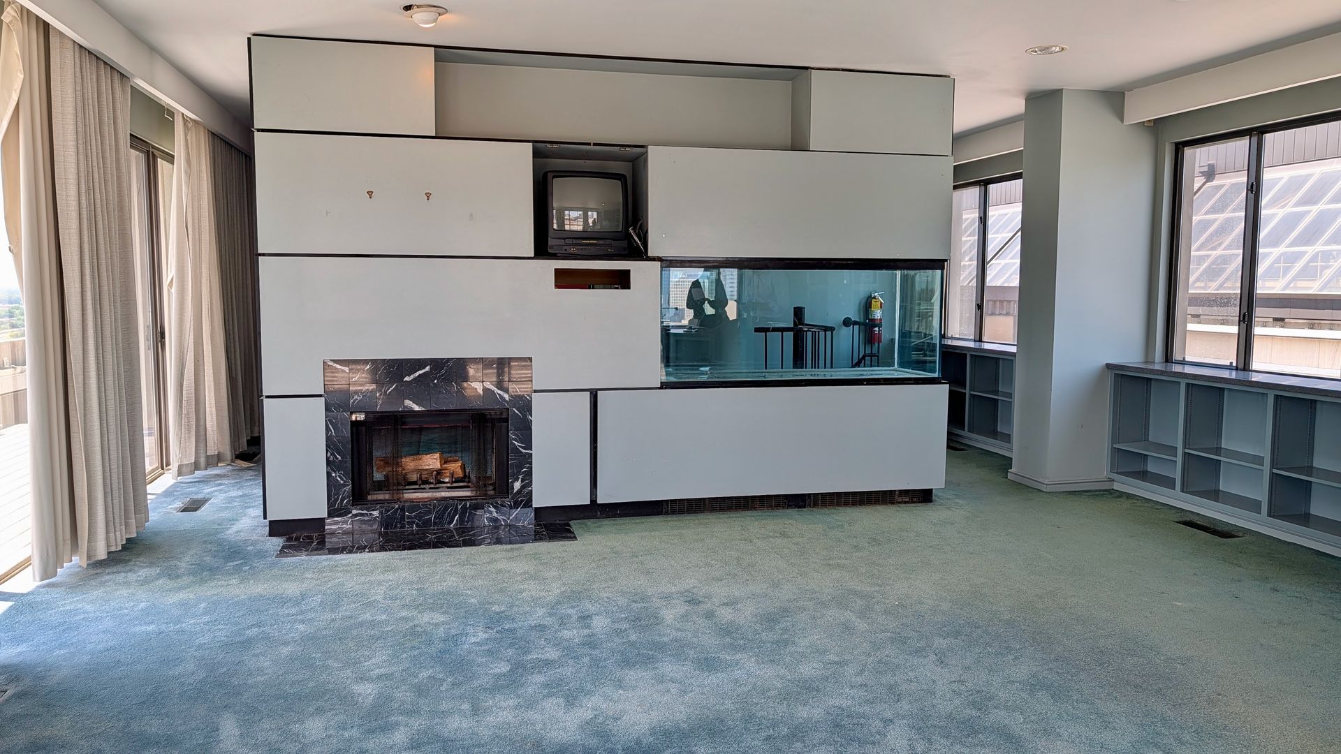 Living room with a light-gray modular wall unit around a small TV above a marble fireplace. A large glass fish tank is built into the wall; floor-to-ceiling windows with beige curtains and a blue carpet.