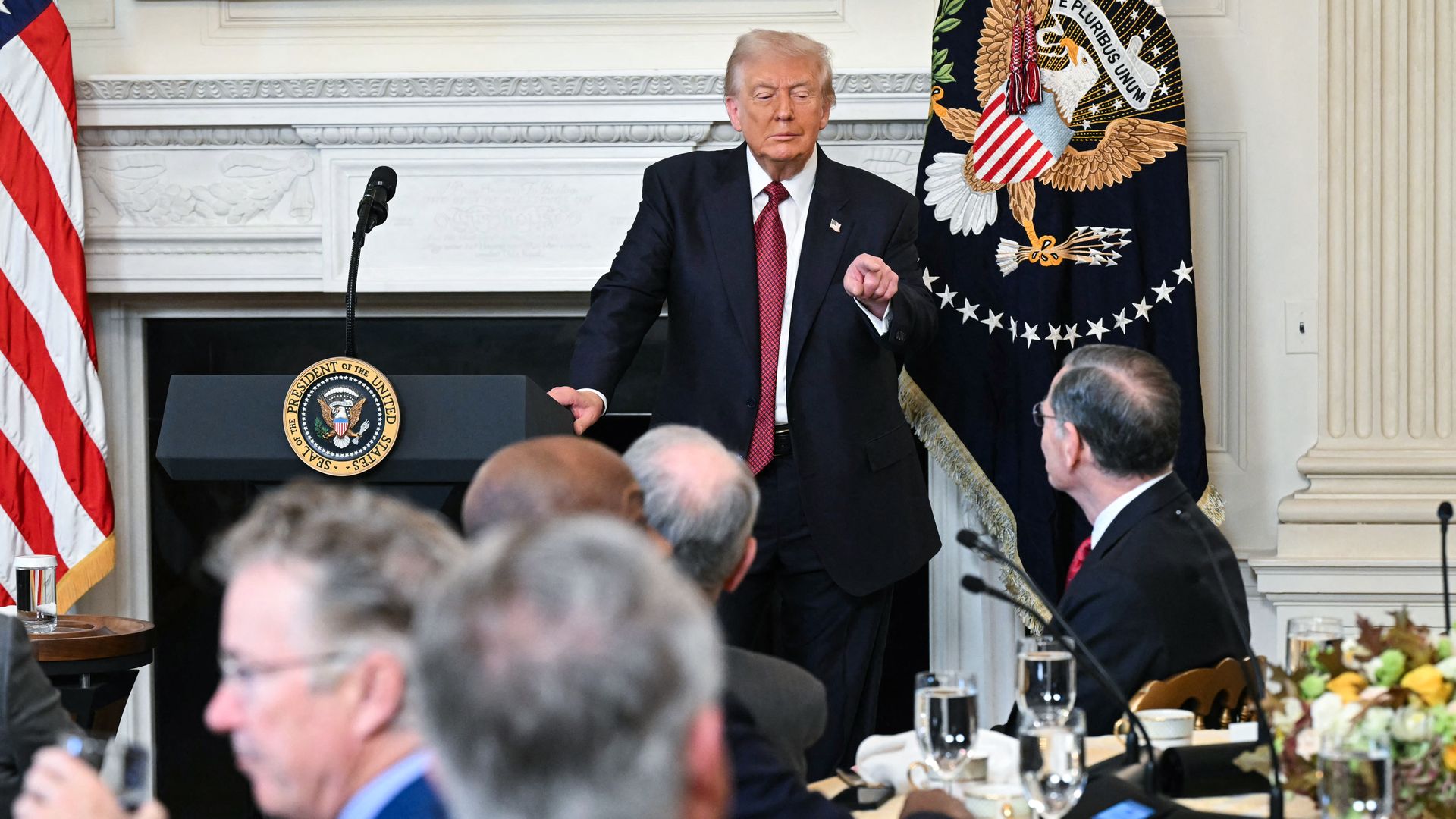 Trump stands with one hand on a podium with a microphone as he points at a man in a crowd of people sitting at tables with the other. He is wearing a dark suit, a red tie, a white collared shirt and an American flag pin.