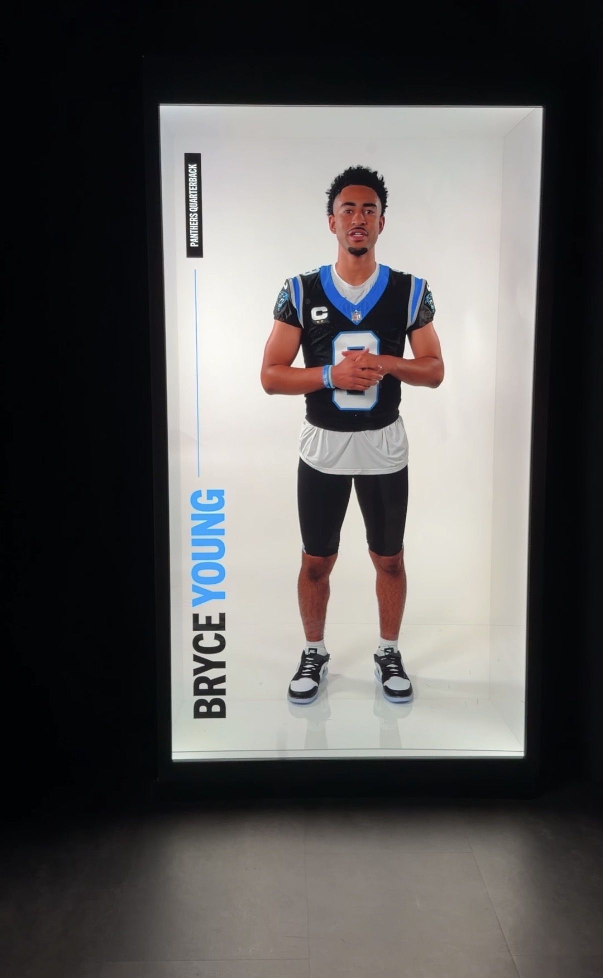 Life-size digital display of NFL QB Bryce Young in a Carolina Panthers black jersey with blue trim and a captain patch, standing with hands clasped.
