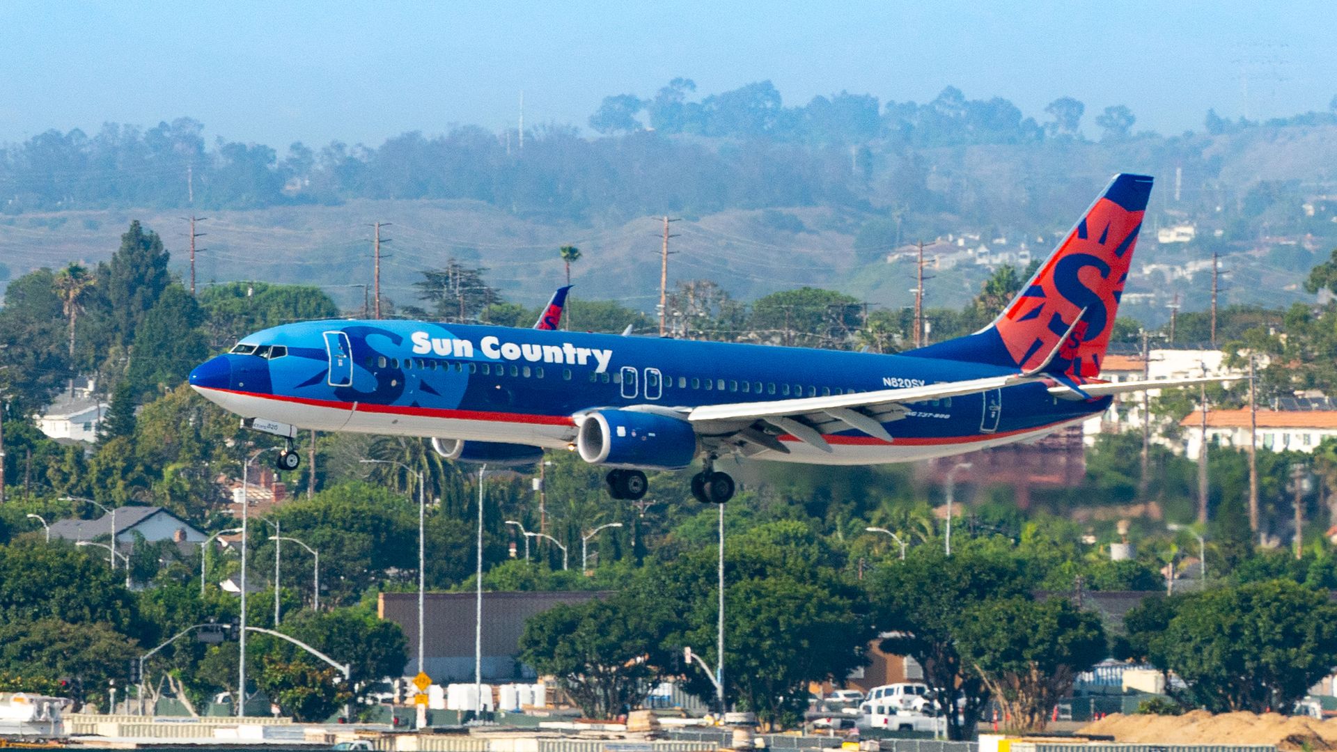 A blue and orange Sun Country plane is landing with trees in the background. 