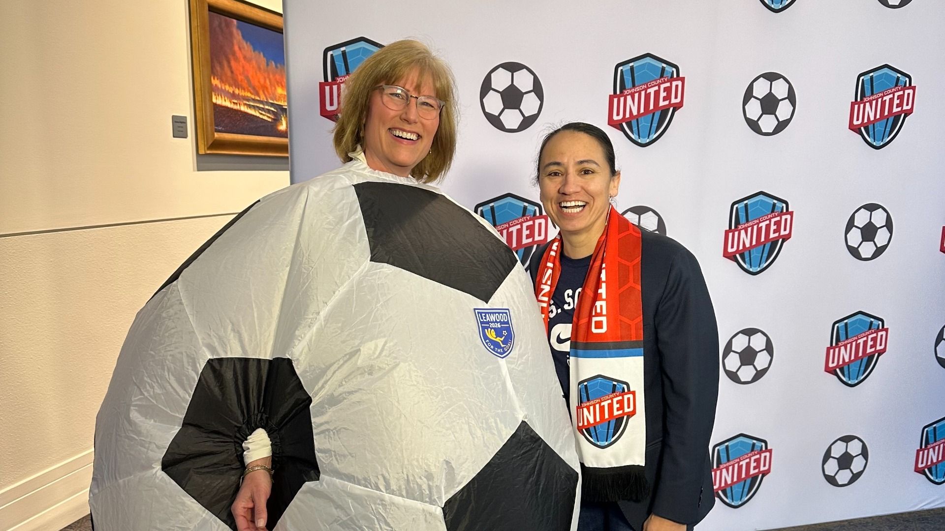 Two women smile at a soccer event; one wears a large gray and black soccer ball costume with a Leawood 2026 patch, the other sports a red United scarf, against a backdrop of soccer logos.