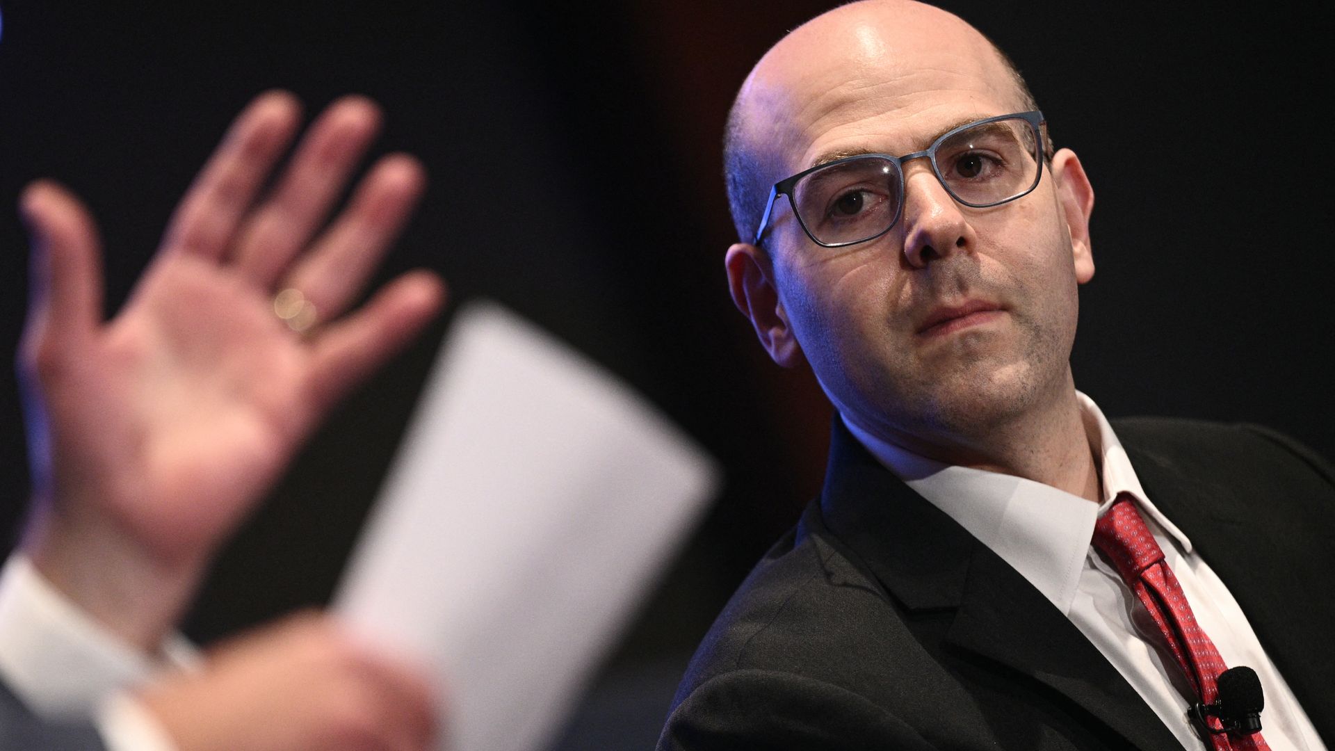 Council of Economic Advisors Stephen Miran in glasses, a black suit, white shirt, and red tie listens attentively; a blurred hand in the foreground holds a paper, gesturing during a discussion or interview.