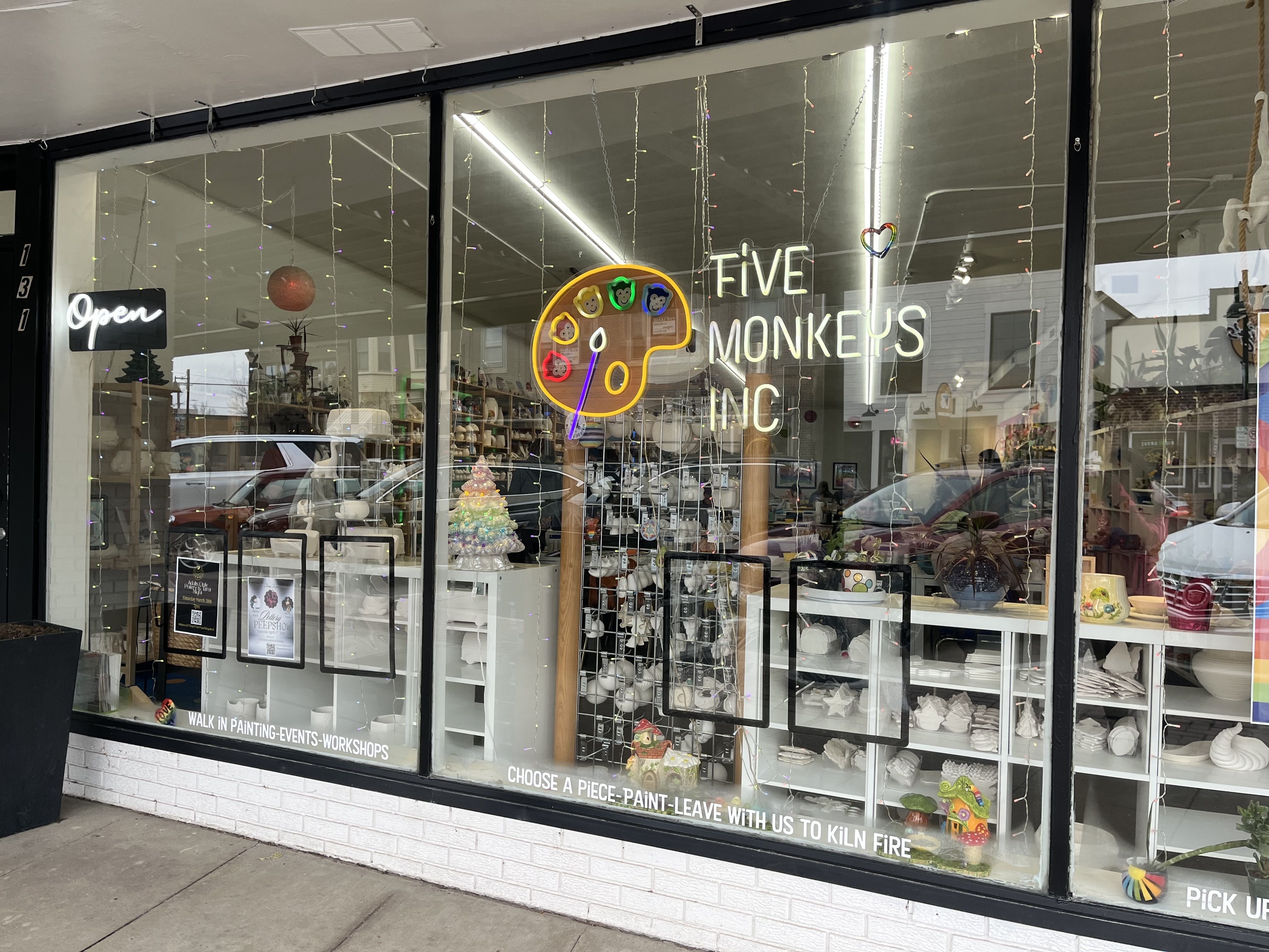 Shop window of Five Monkeys Inc art studio with neon sign shaped like a yellow palette and text, displaying ceramic pieces and a rainbow heart decoration inside.