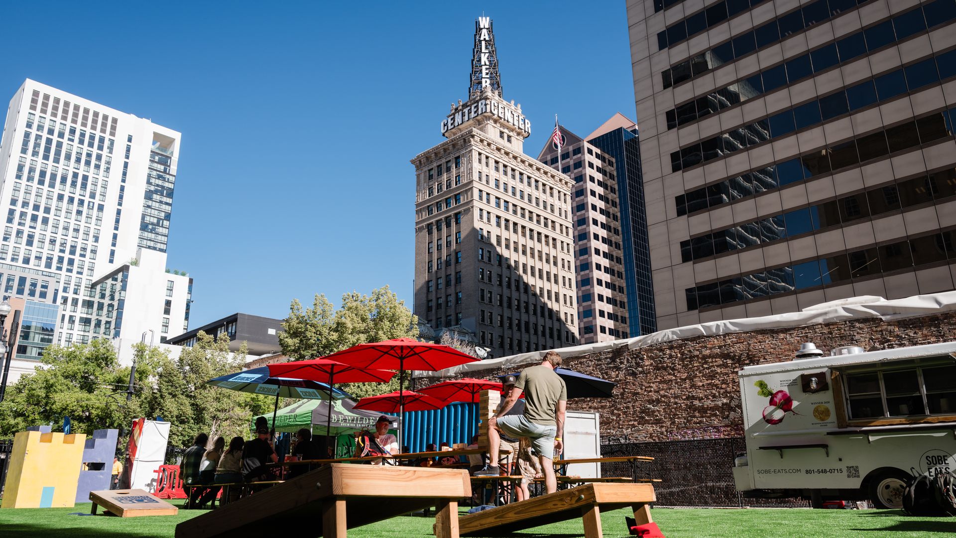 Downtown skyline with a tall historic building labeled Walker Center atop a spire, beside modern glass towers; foreground shows red umbrellas, outdoor tables, and a food truck.