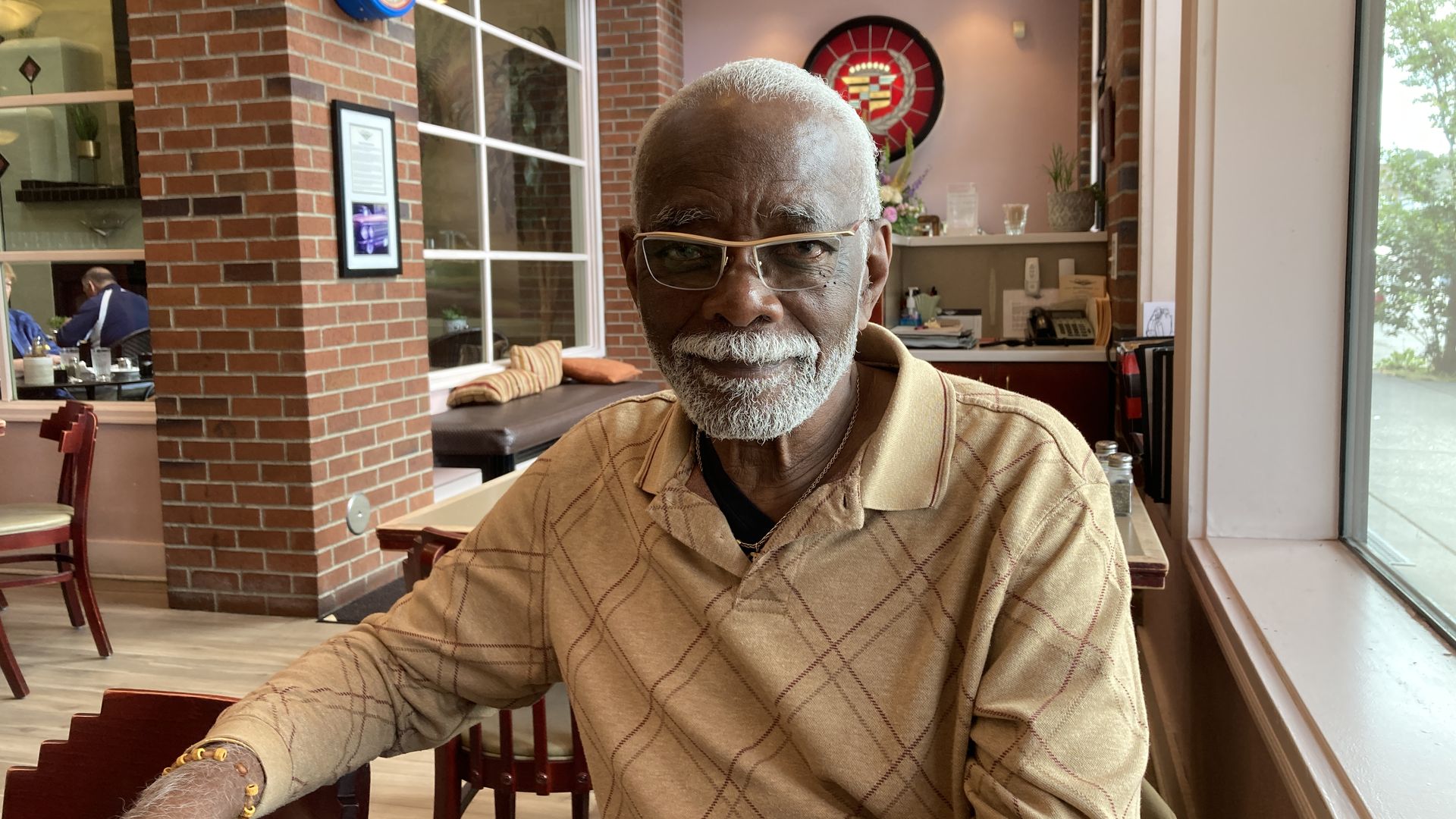 A man with a grey beard and mustache, wearing glasses, and sitting at a restaurant table, looks at the camera.