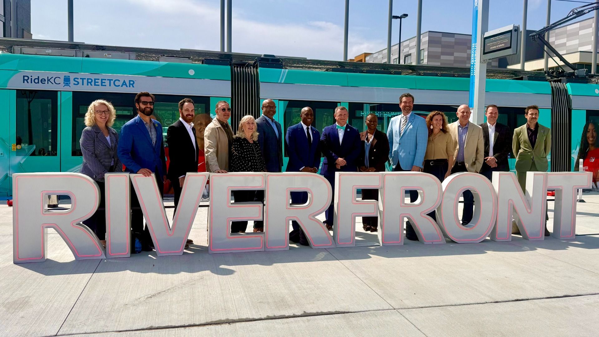 A diverse group of professionals in smart casual attire pose in front of a teal RideKC Streetcar. They stand behind oversized white letters with pink edges spelling a large word on the pavement.