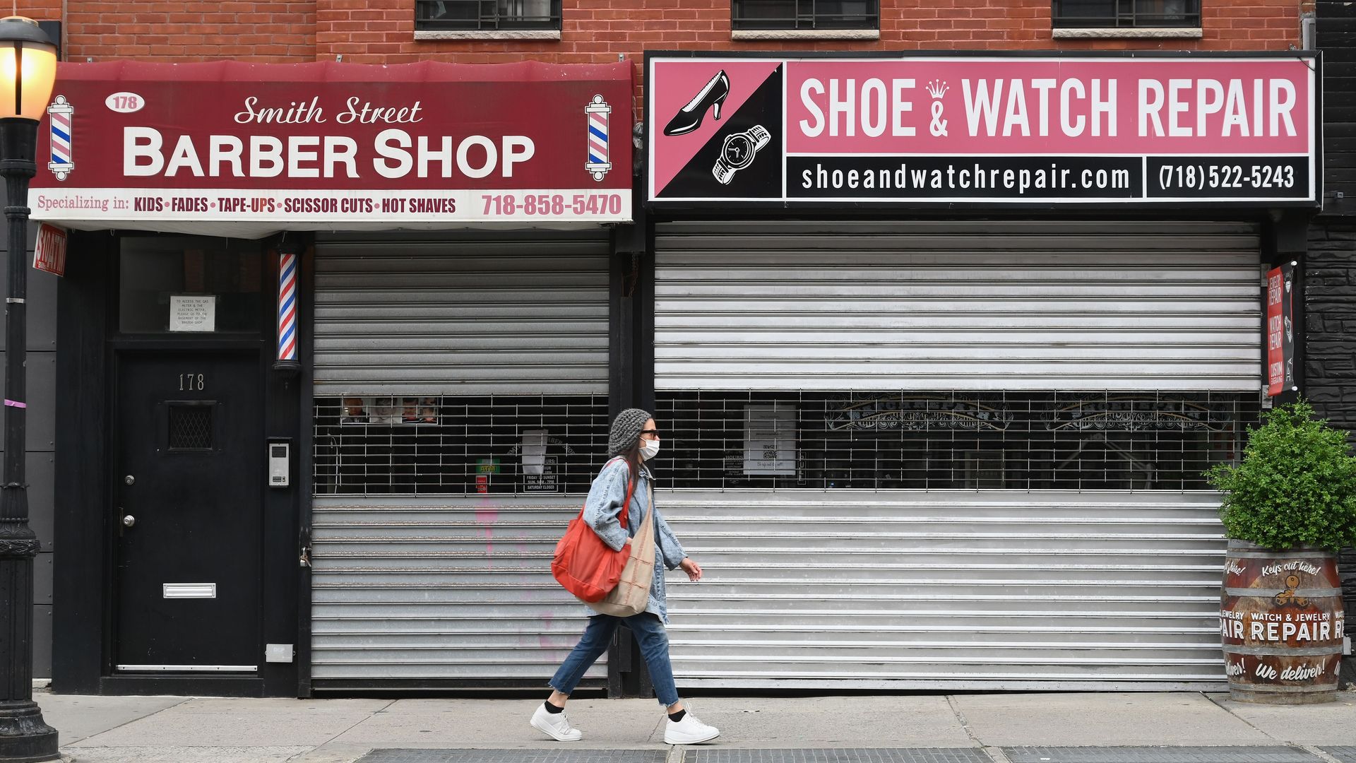 A person in a mask walk by a closed barber shop and closed watch repair shop.