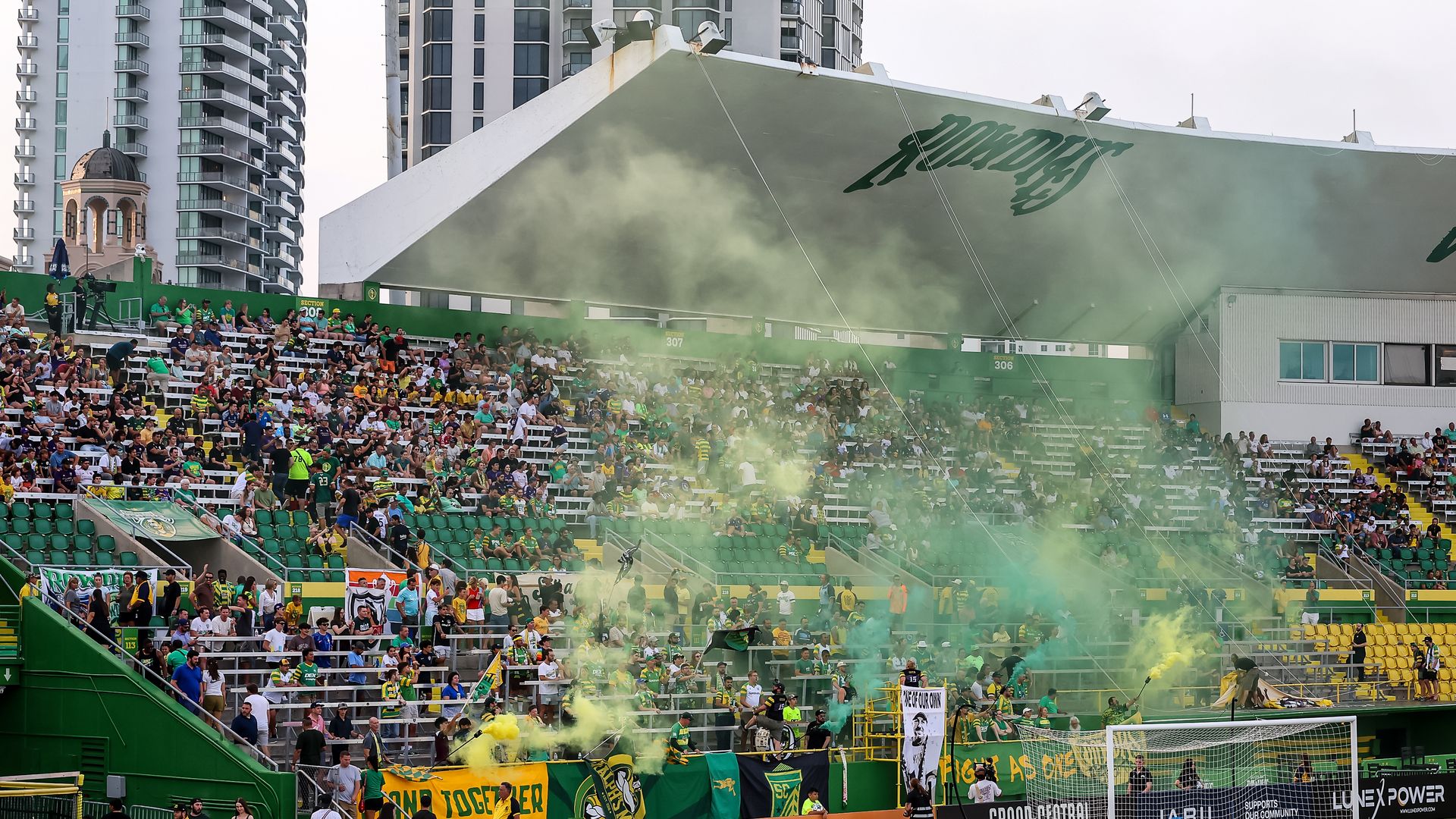 Busy soccer stadium filled with green-clad fans; green and yellow seats, banners, and smoke from flares near the goal. Urban skyline in background beneath a white canopy.