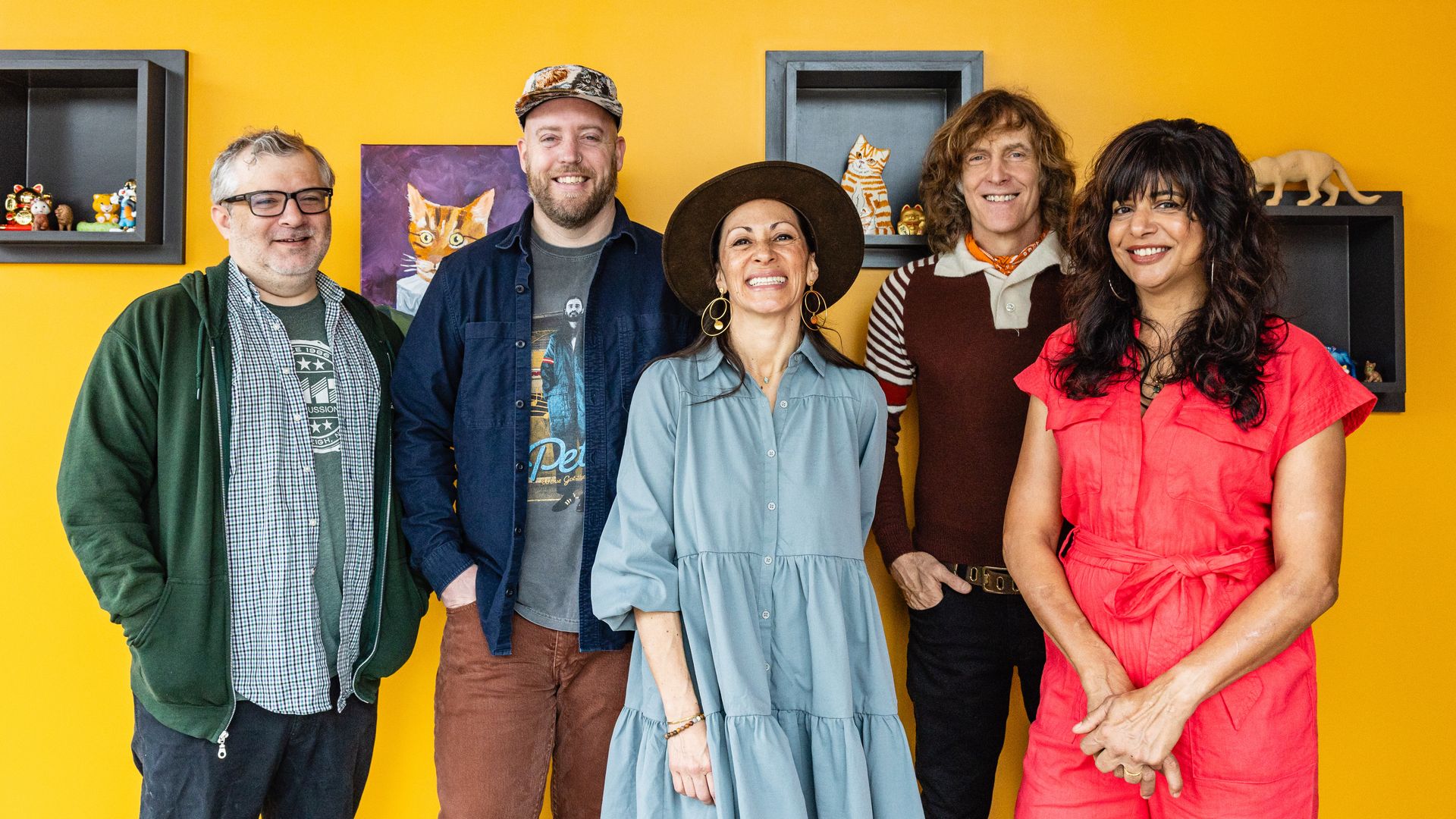 Five smiling adults standing in front of a bright yellow wall with shelves holding cat figurines and paintings. They wear casual, colorful clothing and appear cheerful.