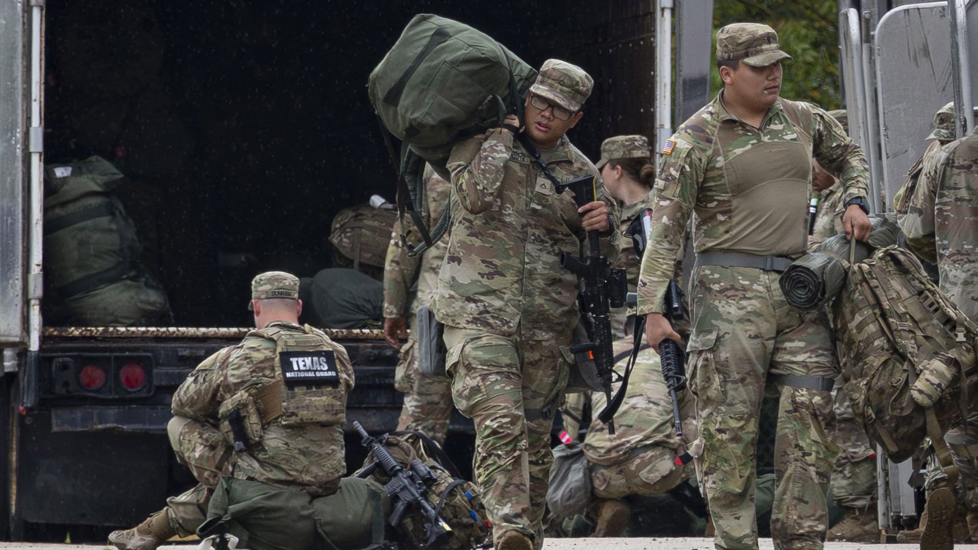 Camouflage-uniformed, Texas National Guard, wearing caps and carrying Army bags and guns, walk past a truck full of Army bags as a fair-skinned member sits on a bag with his back to the camera. The words "Texas National Guard" are displayed in white writing with a black background on his back.