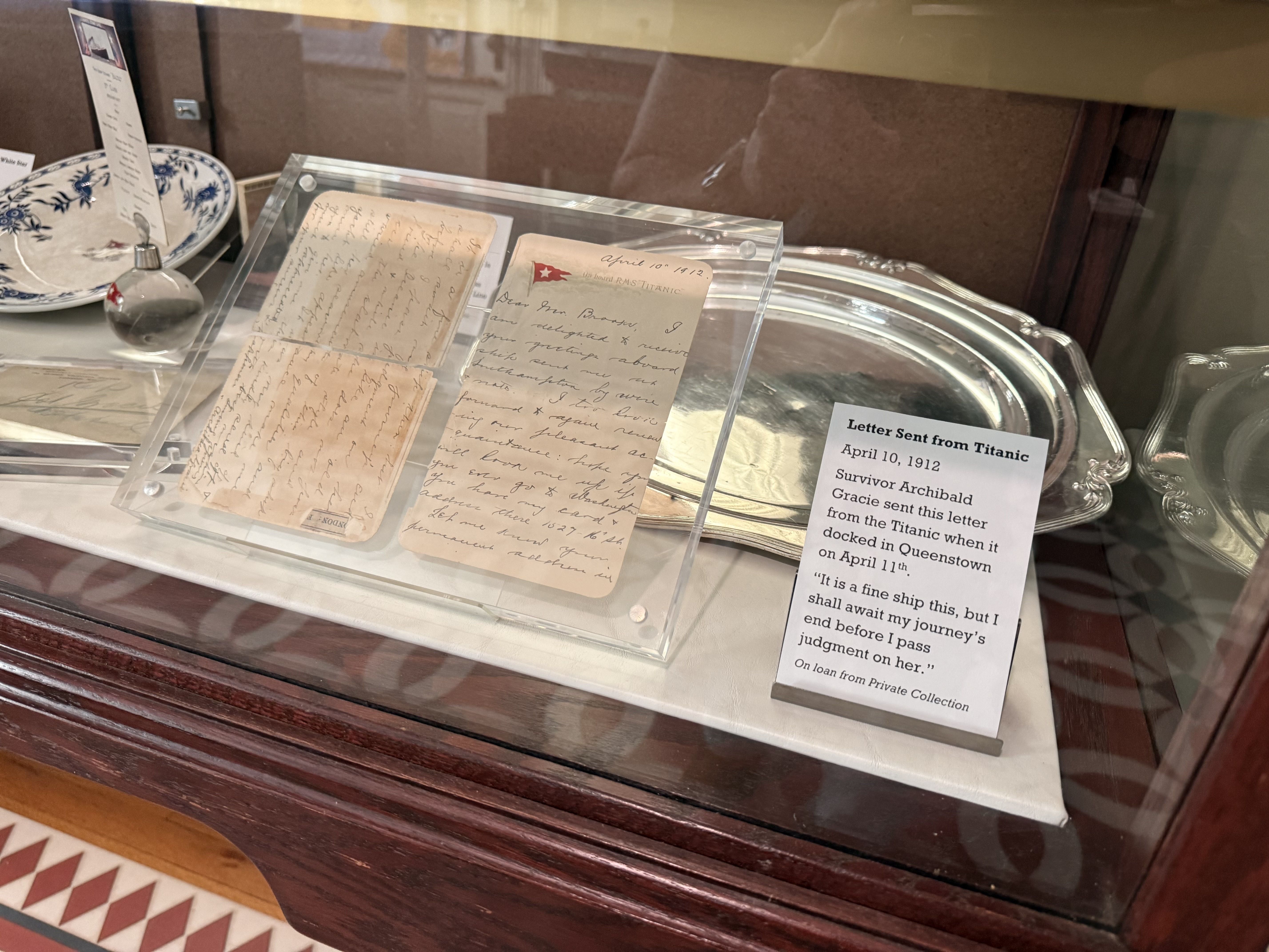 Glass display case showing two aged handwritten letters mounted in clear sleeves, a small red flag atop one page, with a silver tray in the back. A label reads "Letter Sent from Titanic" (1912).