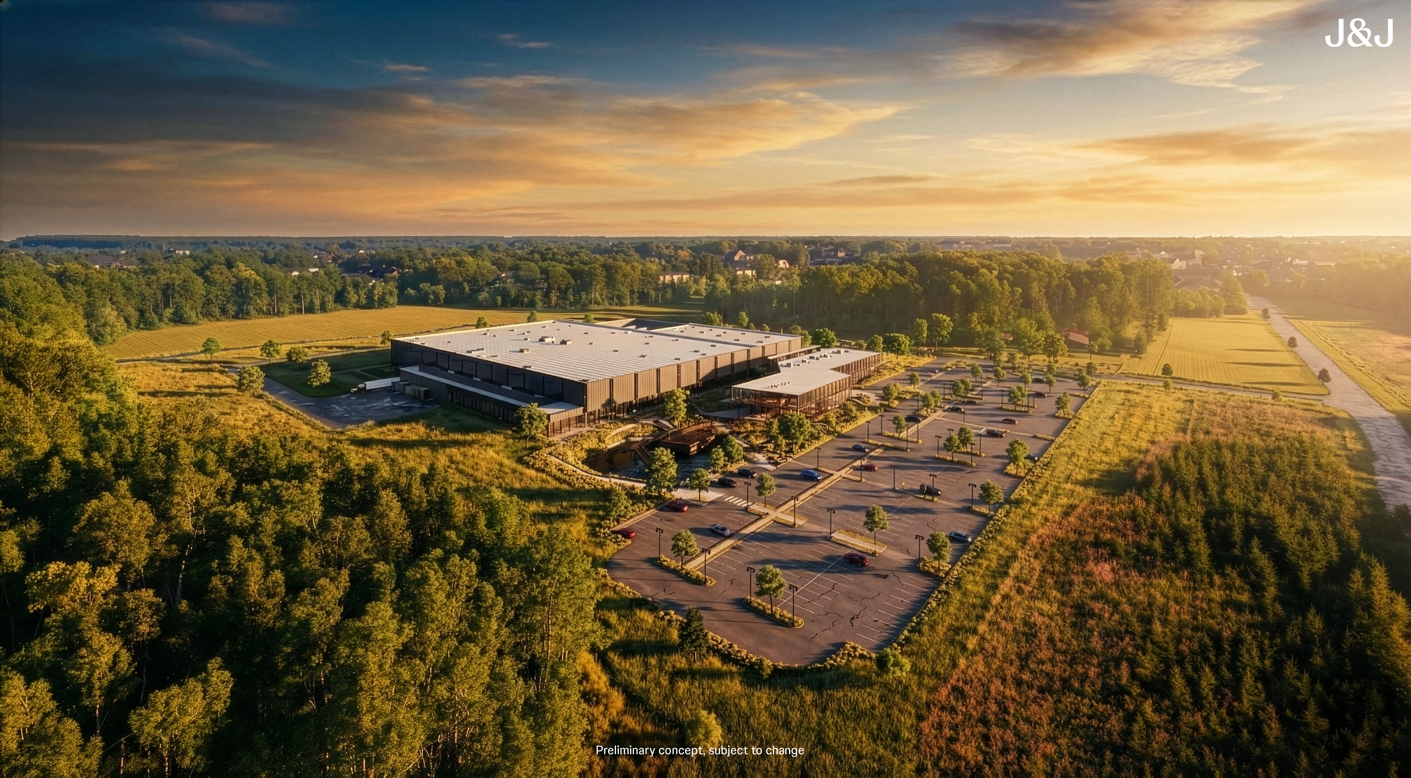 A large modern warehouse with a gray roof surrounded by parking lots and green trees during a golden sunset, with fields and a distant neighborhood in the background.