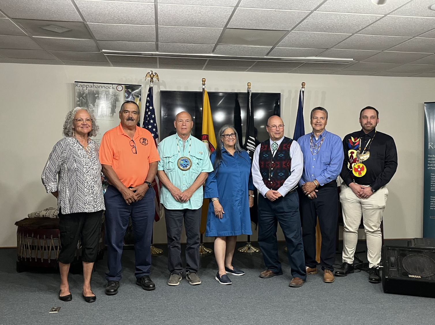 Seven people stand in a line indoors, dressed from casual to ceremonial. Behind them are flags, a large screen, and a wall display labeled Rappahannock.