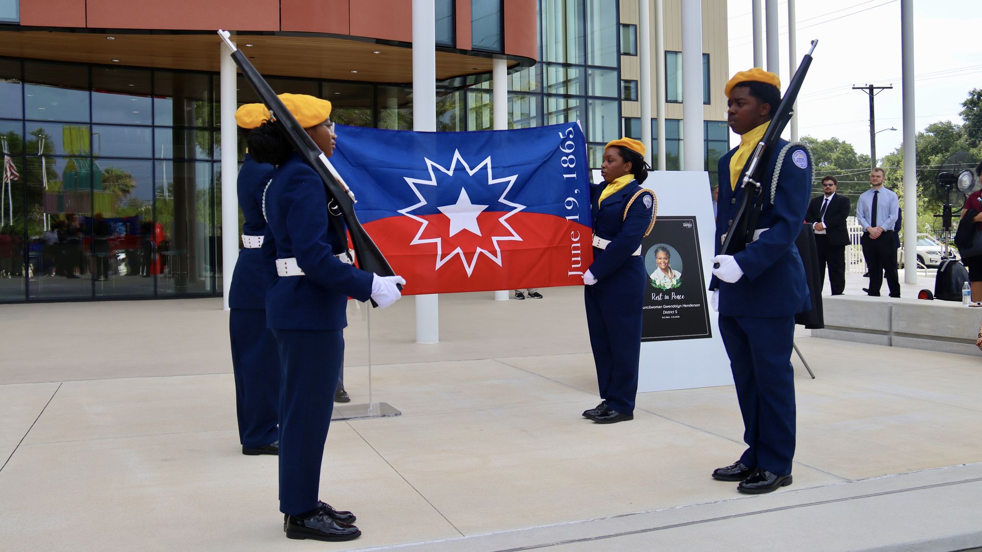 Four people in navy uniforms and yellow berets hold the red, blue and white Juneteenth flag in front of a windowed building.
