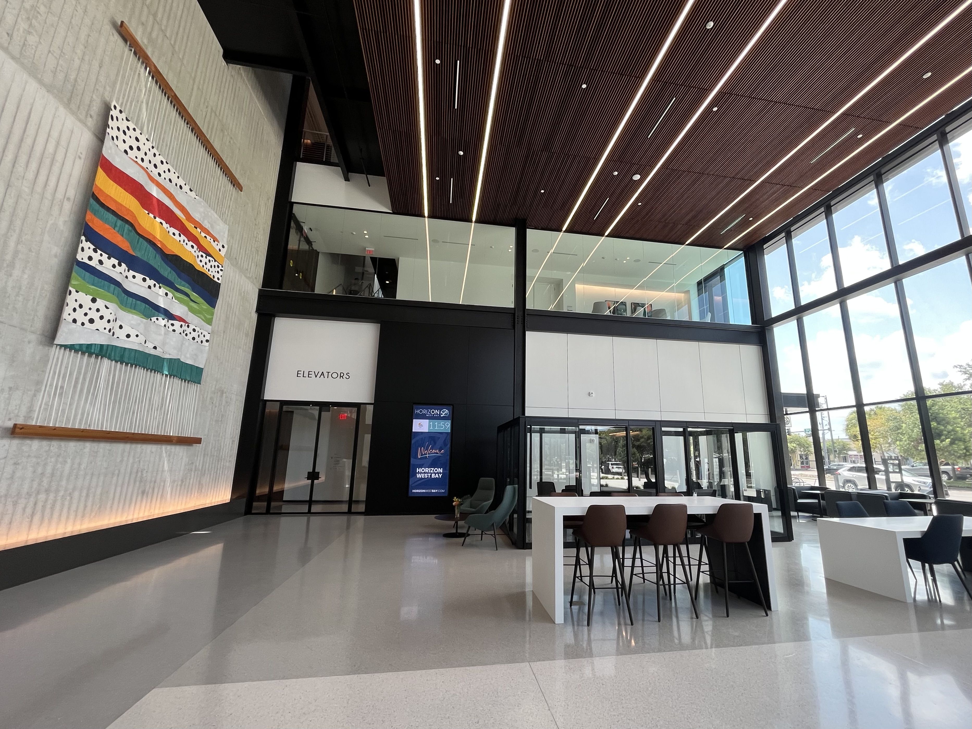 Modern lobby with floor-to-ceiling glass on the right, a dark wood slat ceiling with linear lights, colorful striped wall hanging on the left, and white tables with brown chairs near the elevators.