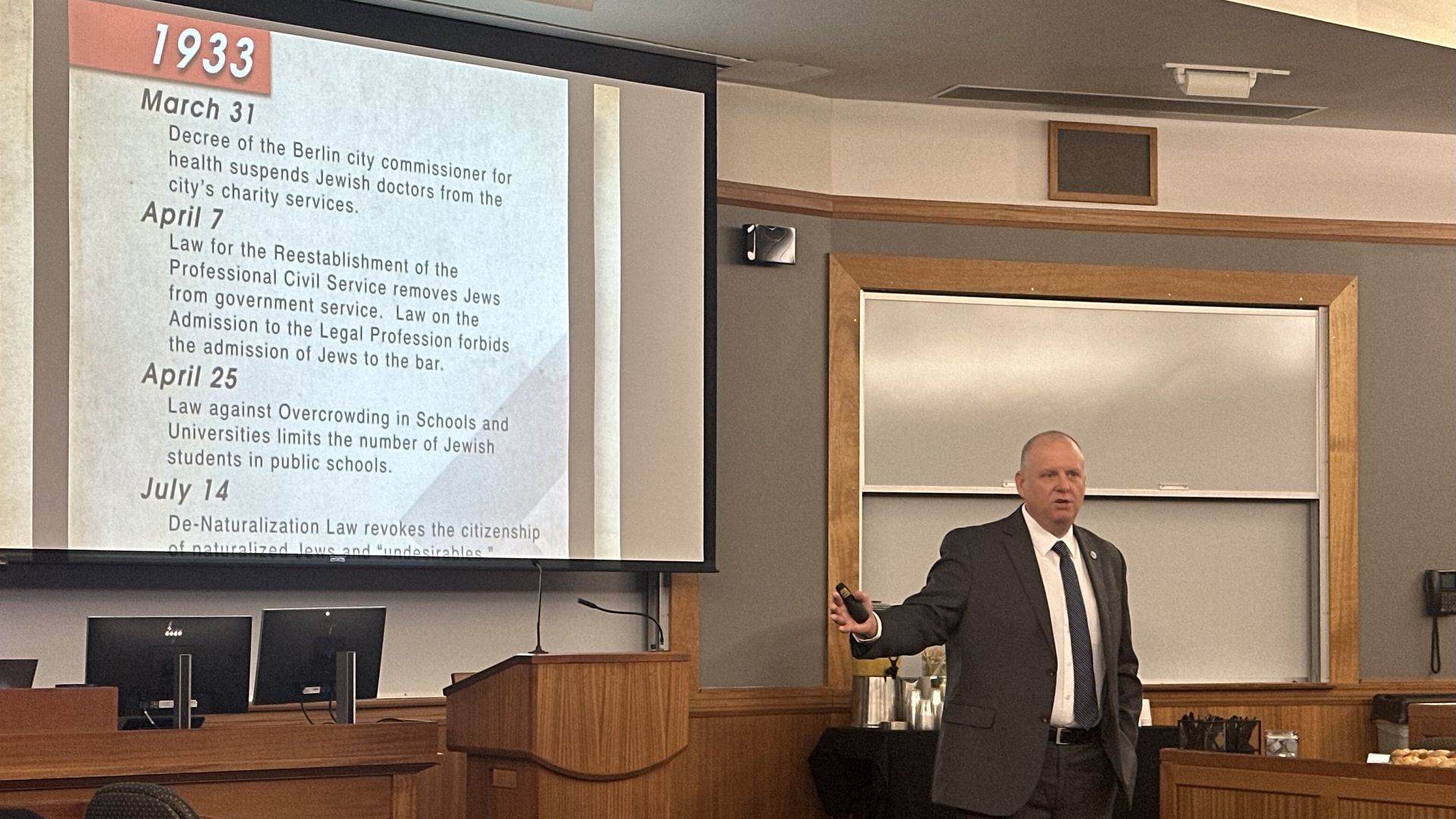 A man in a suit and tie presents in front of a timeline of antisemitic policies in Germany in 1933, during the rise of the Nazi regime. 