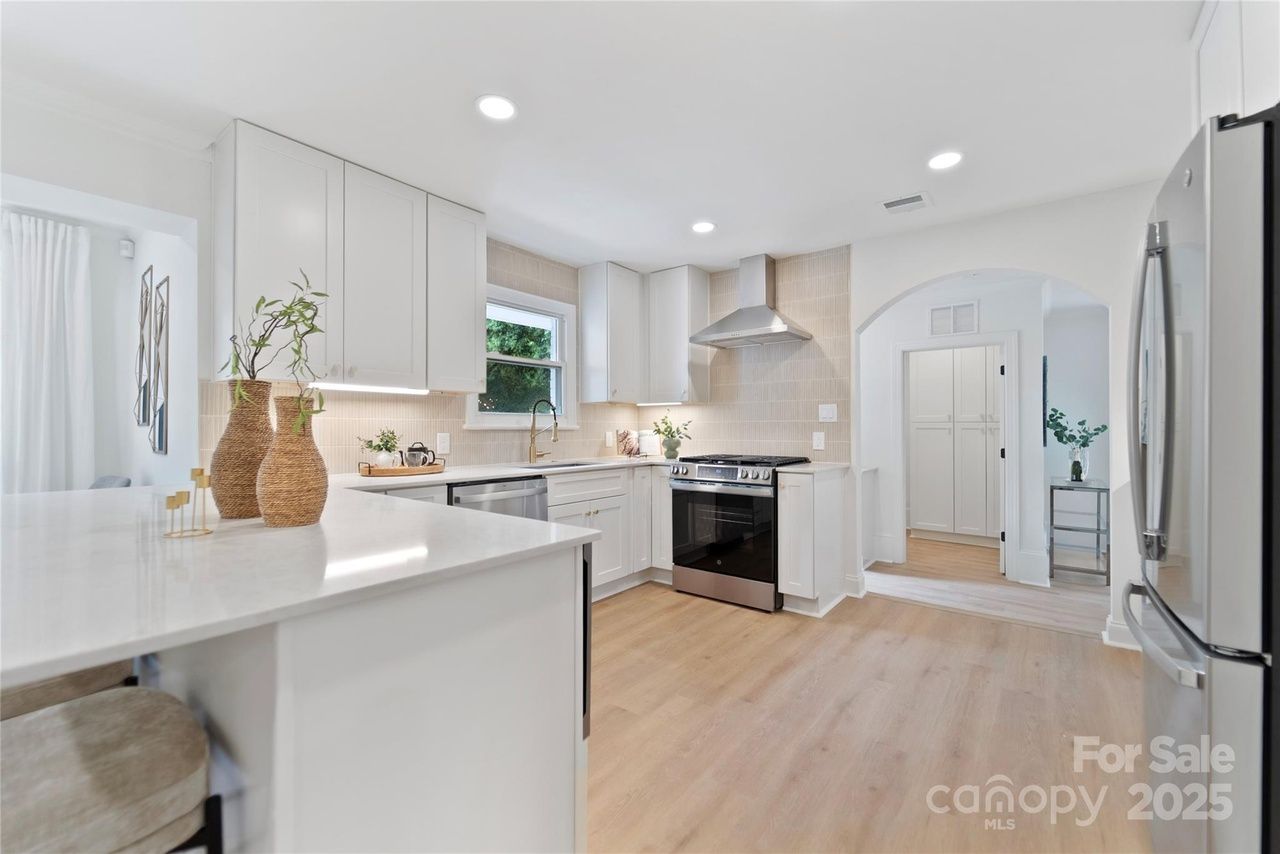 Bright modern kitchen with white cabinets, beige tiled backsplash, stainless steel appliances, light wood floors, granite countertop island with woven vases, and arched doorway leading to another room.