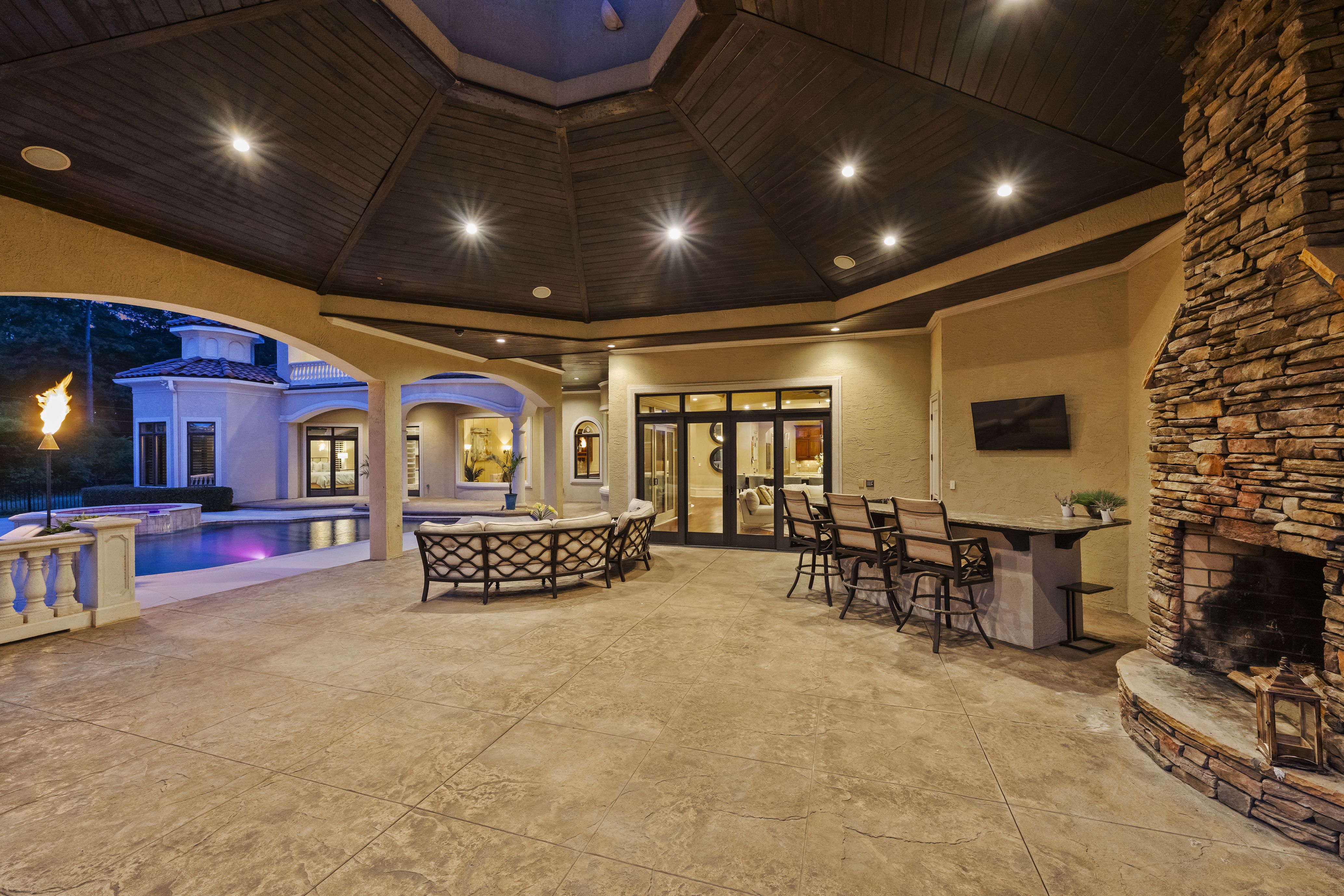 Evening view of a covered outdoor patio with a dark wooden octagonal ceiling, beige walls, a stone fireplace, glass doors, a lit torch by a pool, and wicker seating.