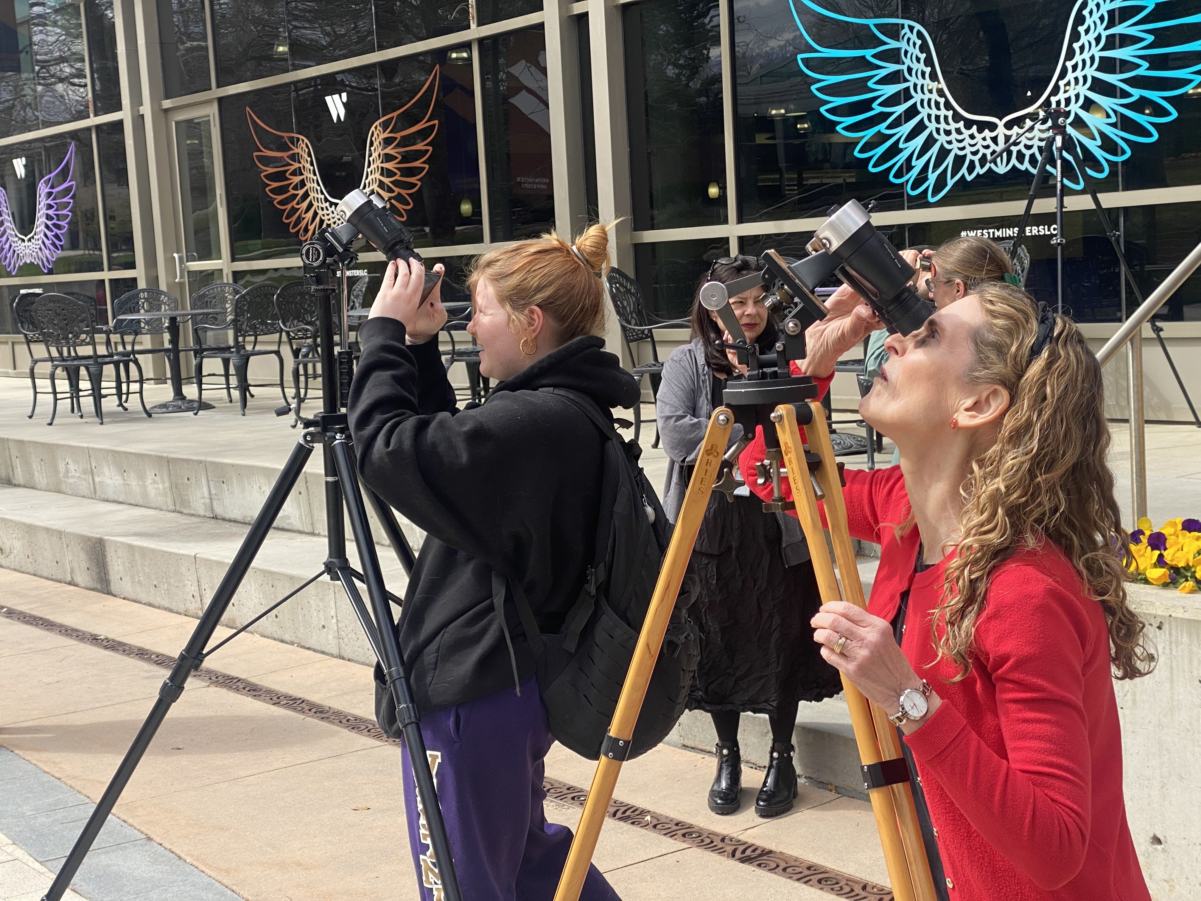 People look at the sky through binoculars in front of a cafe.
