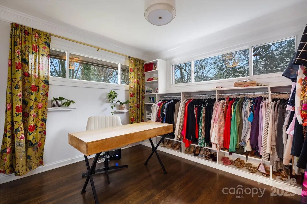 Bright dressing room with wooden floor, yellow floral curtains, white walls, a wooden table with metal legs, white chair, hanging clothes of various colors, and small plants on wall shelves.