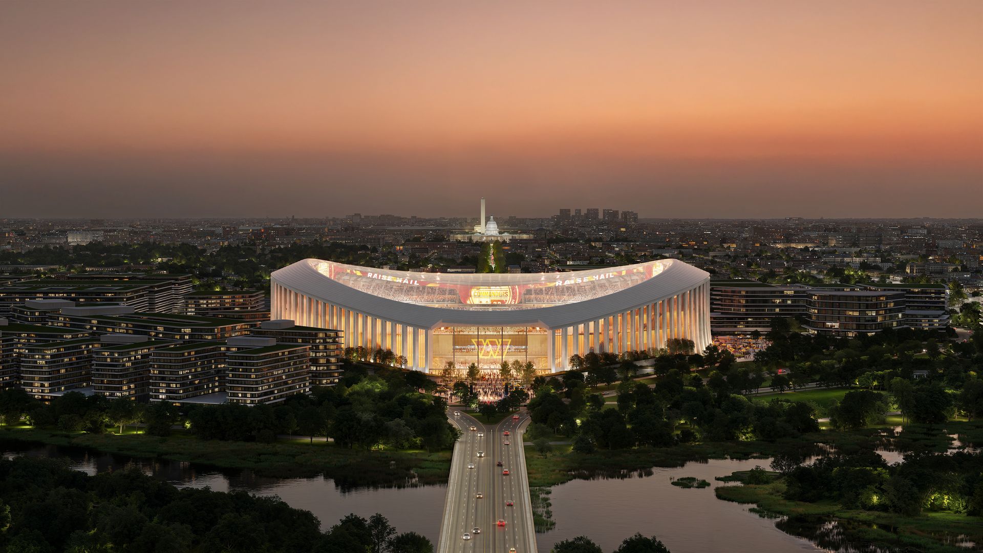 A modern, illuminated stadium at sunset with a visible highway leading to it, surrounded by greenery and buildings, with a distant city skyline featuring a domed white building and a tall monument.