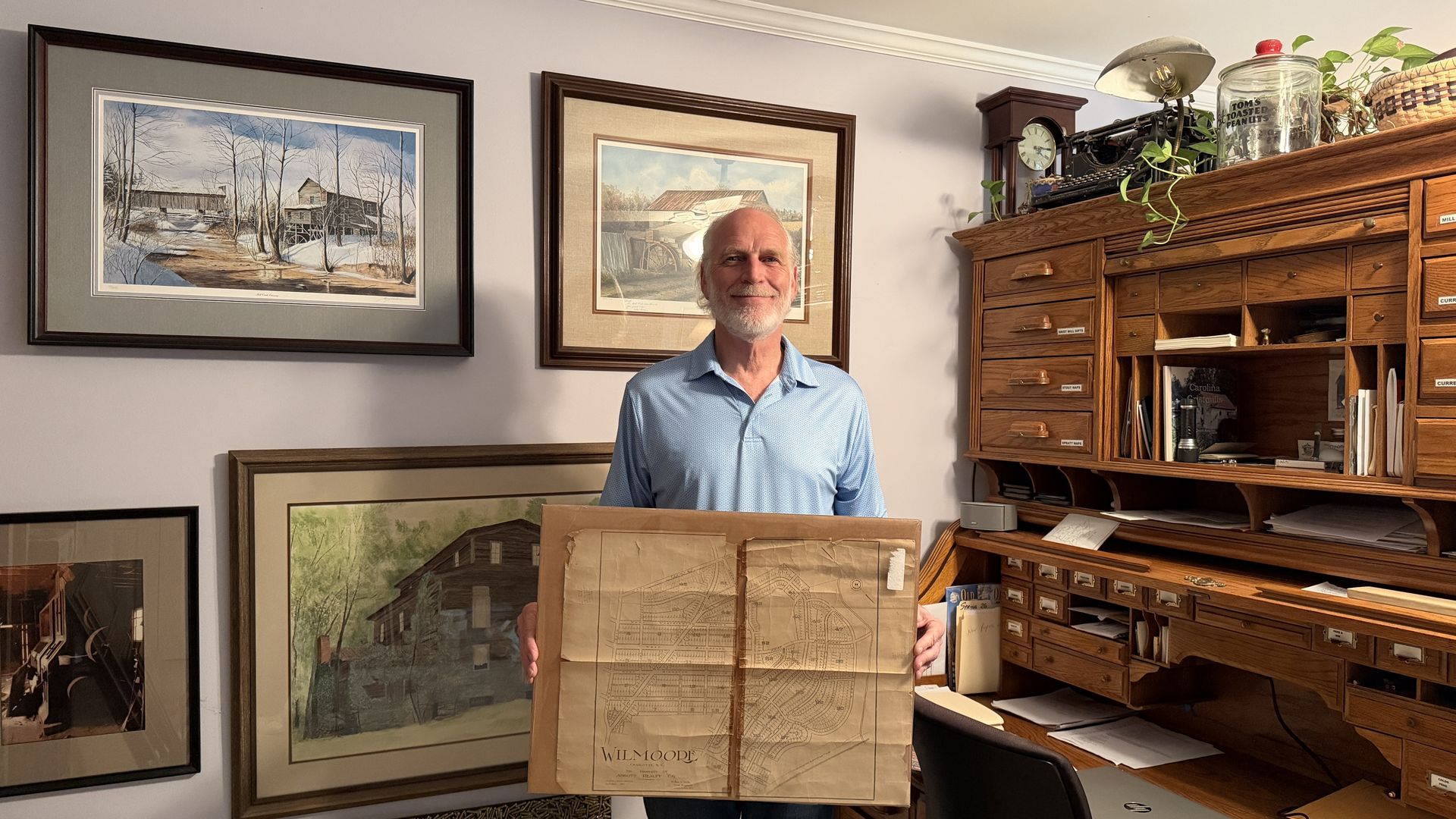 Smiling older man in a light blue polo stands behind a large cardboard box with a vintage map print. He’s in a wood-paneled office with framed landscape art and a tall, multi-drawer desk.