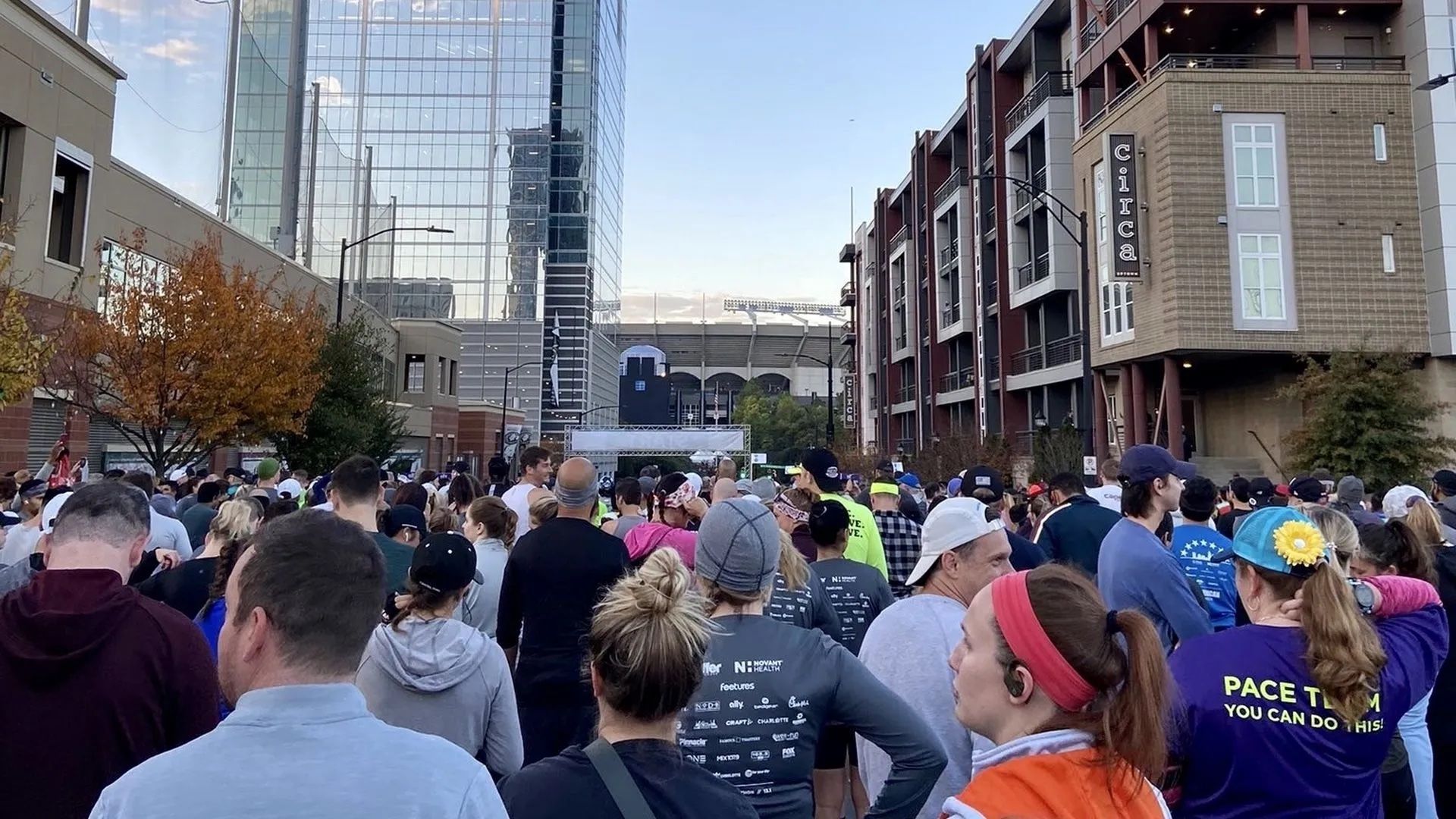 Crowd of people in casual and athletic wear gathered on a city street between tall buildings under a clear sky, appearing to prepare for a run or event near a stadium.